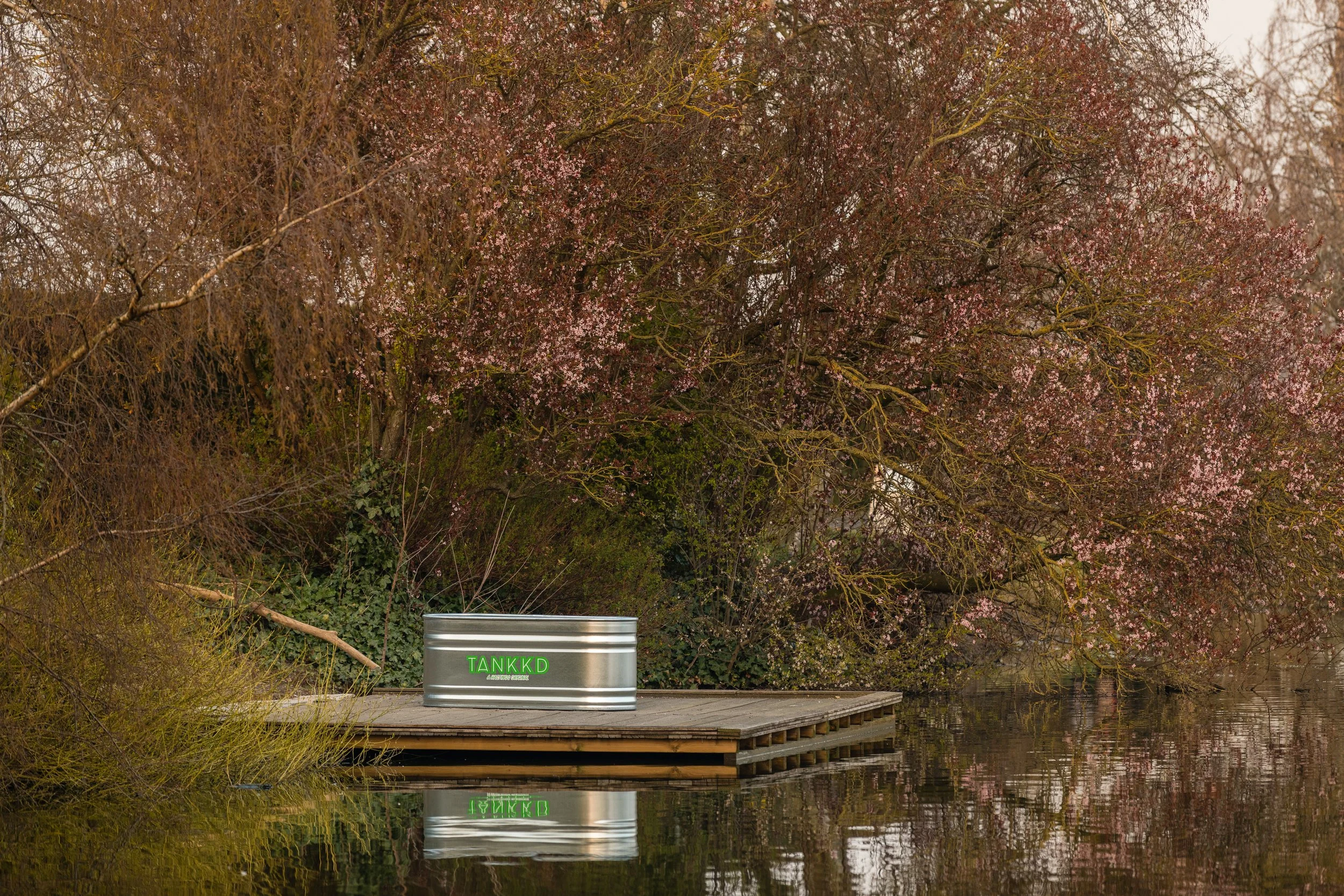 A small Stock Tank on a lake with a metal container labeled 'TANKKD' and surrounded by flowering trees with pink blossoms.