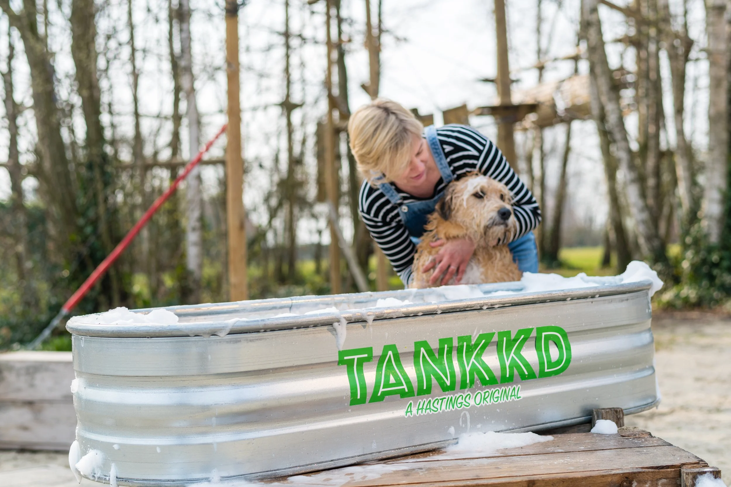 A woman helping a wet dog out of a large Stock Tank labeled "TANKKD, A Hastings Original" outdoors with trees in the background.