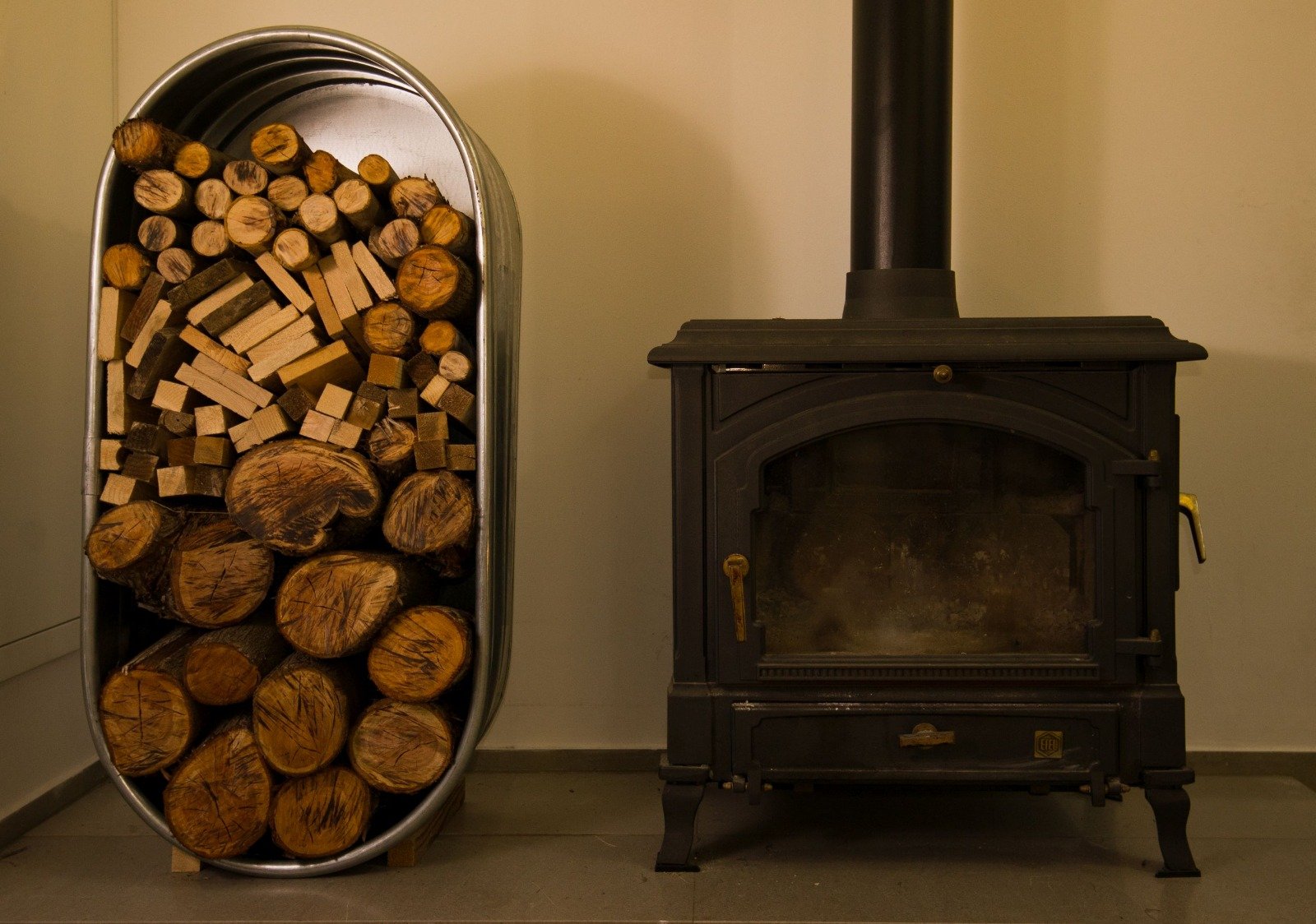 A wood stove next to a metal container filled with firewood logs of different sizes and shapes in a room corner.