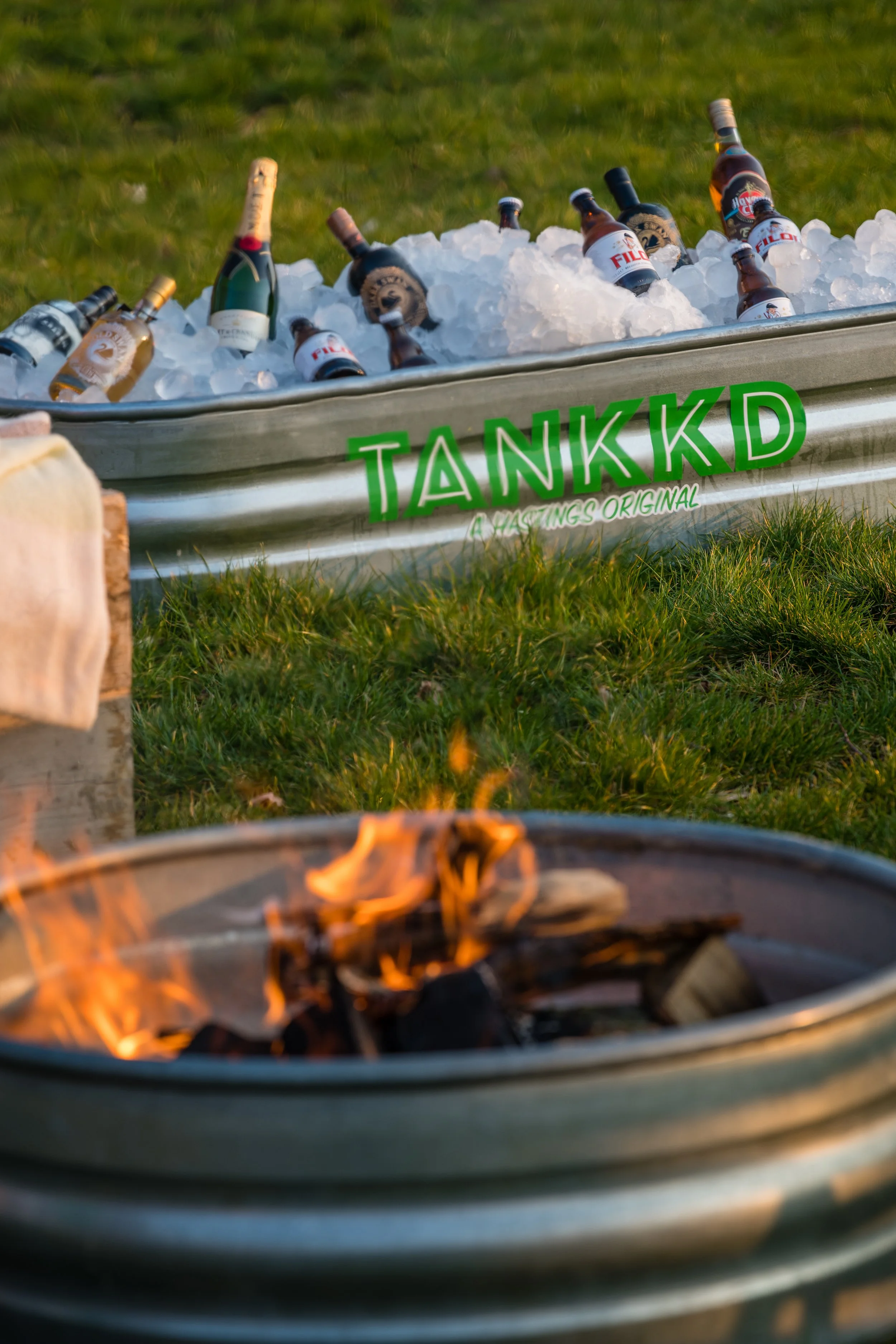 A metal trough labeled 'TANKKD' filled with ice and bottles of alcohol, outdoors on grass, with a small fire in a container in front.