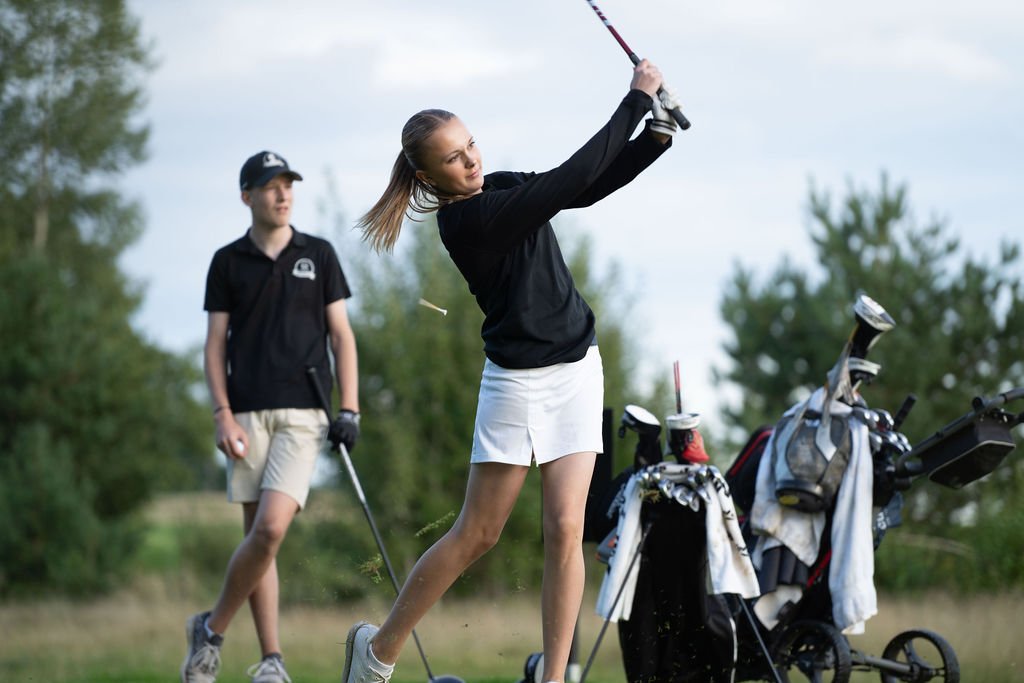 Två personer spelar golf på en grön golfbana, en kvinna svingar en golfklubba och en man står bakom henne. Golfbagar syns i bakgrunden. På Motala Golfklubb.