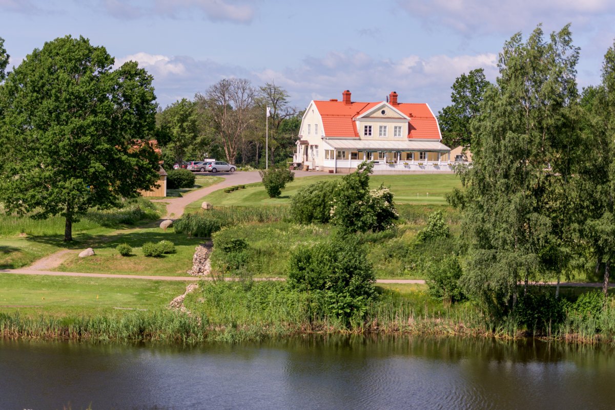 Vit byggnad med rött tak vid en grönskande å, grusstig och parkerade bilar i bakgrunden. Byggnaden är restaurangen The Fairway på Motala Golfklubb.