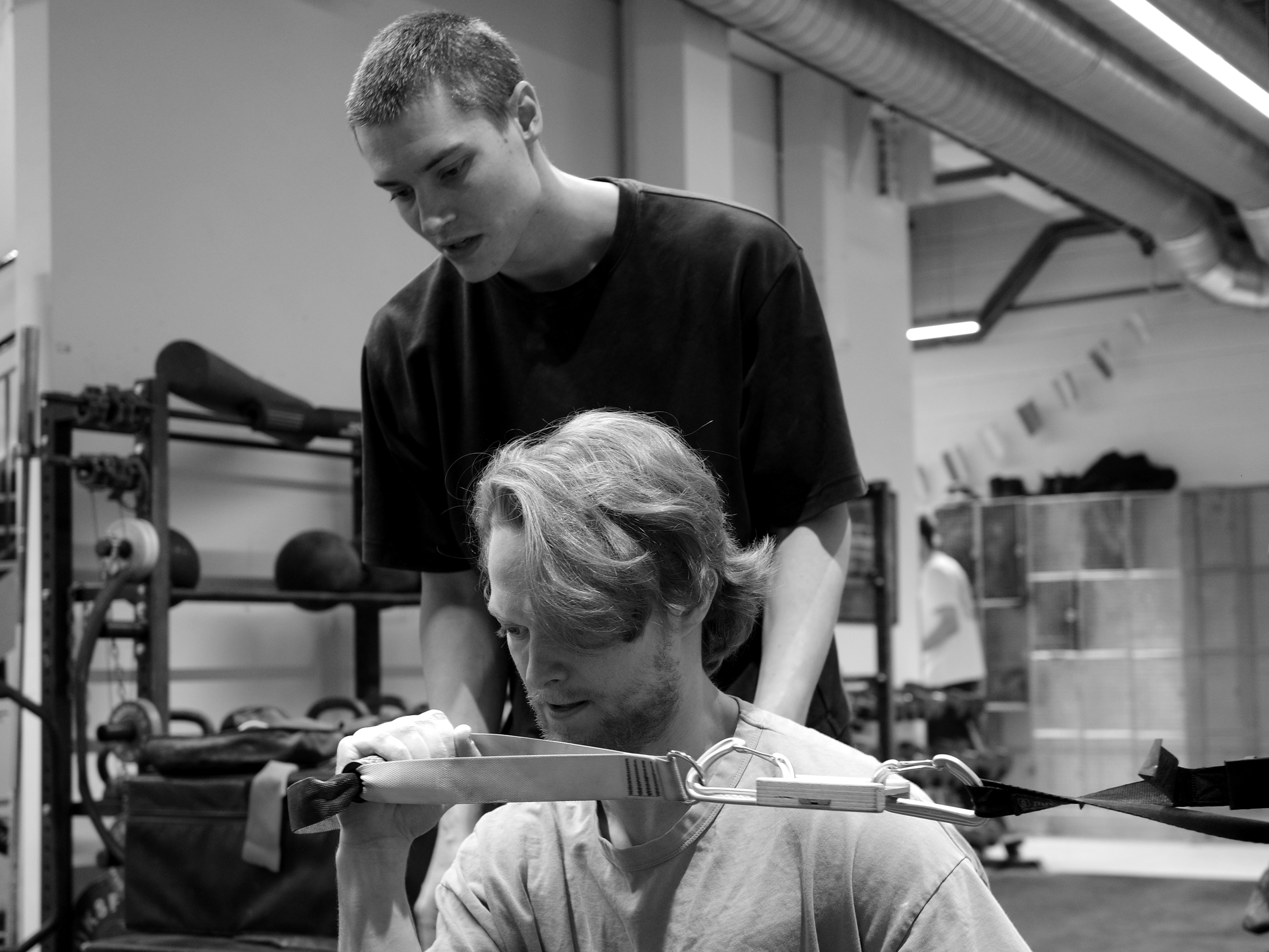 A young man helps an older man with a resistance band exercise at a gym.