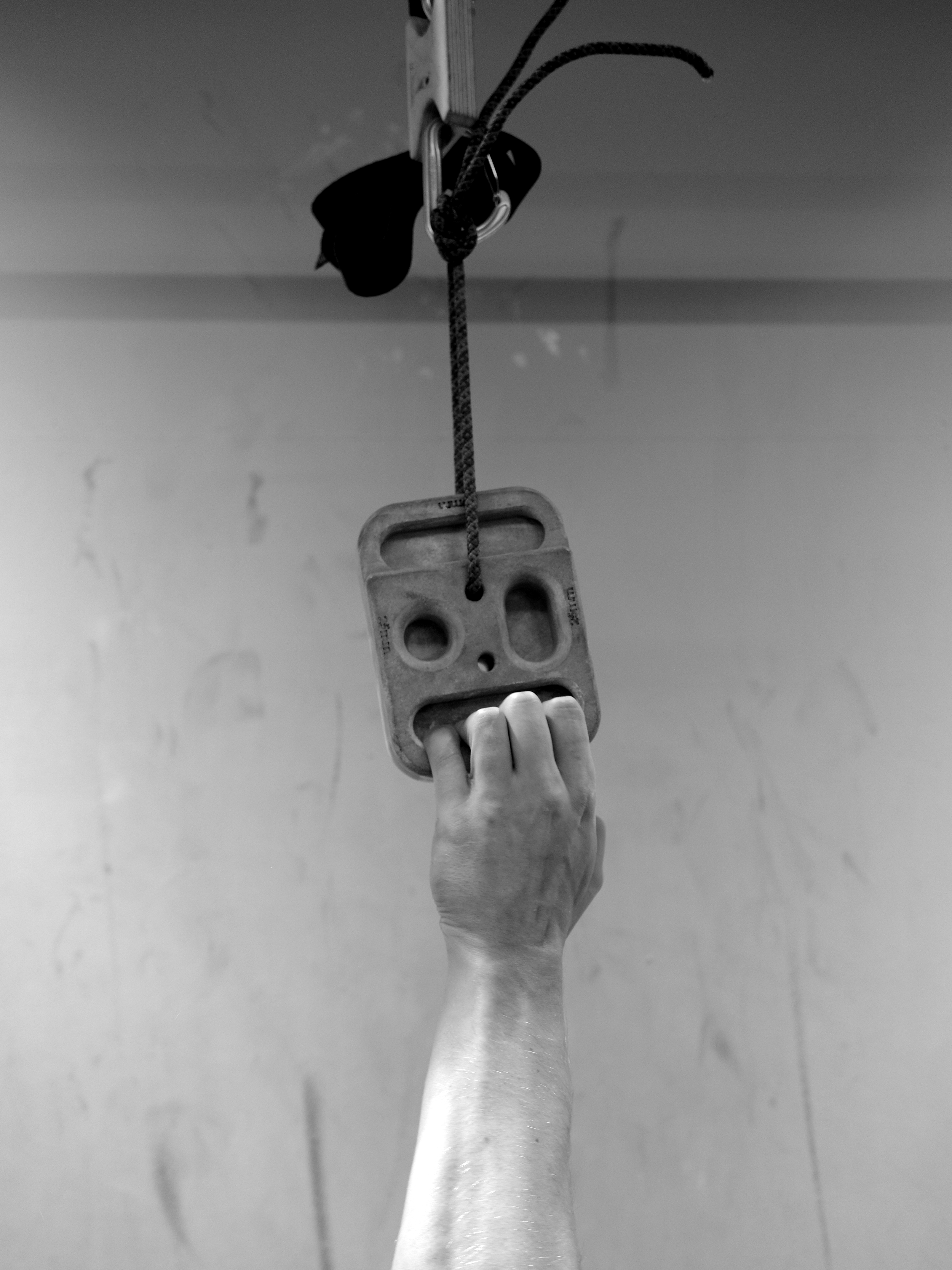 A hand reaching up to grab a training hold on a climbing wall, with a harness and carabiner hanging above.