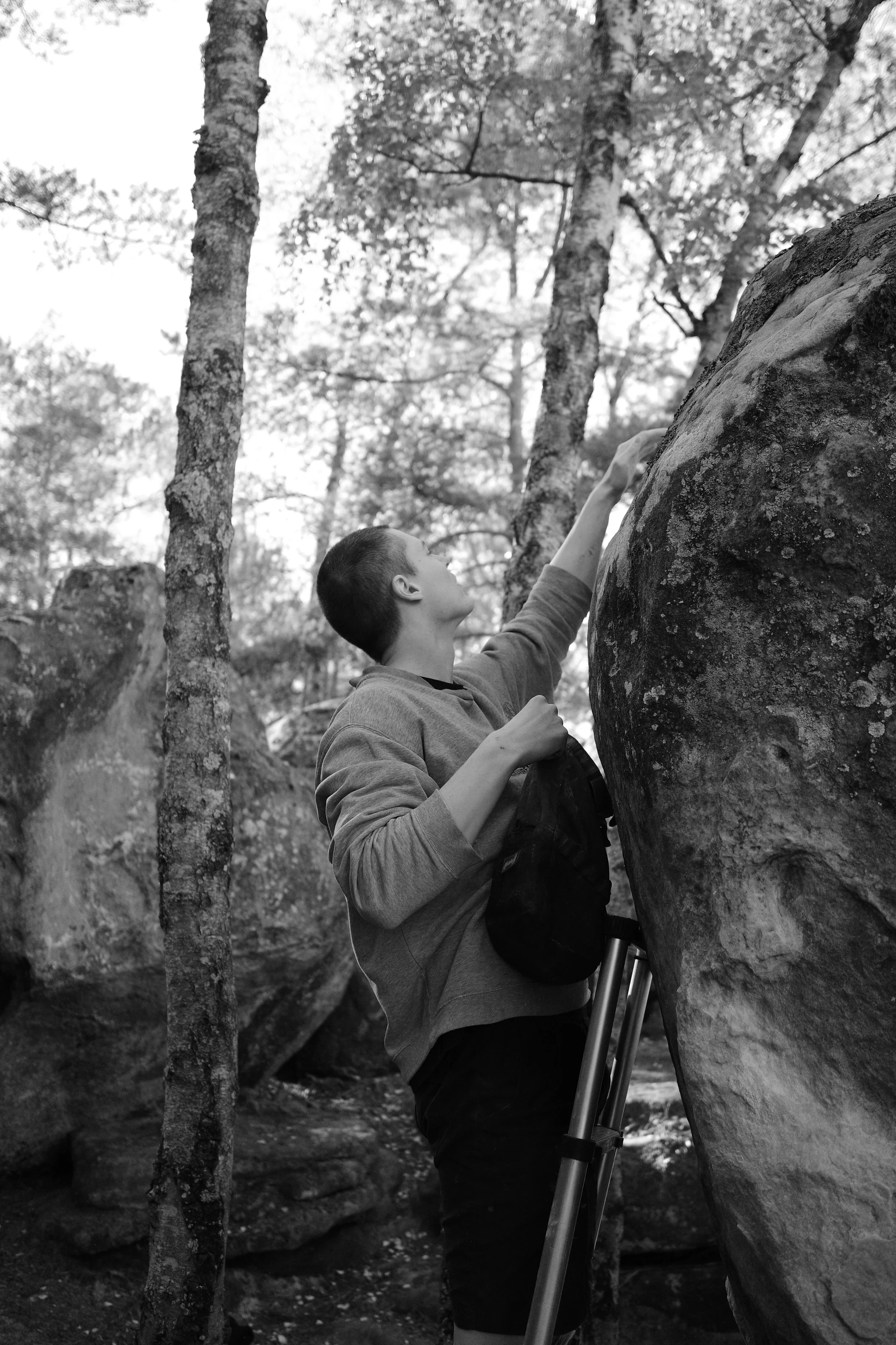 A young man climbing a large boulder in a forest, with trees and rocks in the background, in black and white.