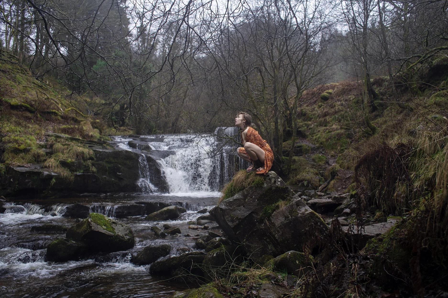 A person with short hair wearing an orange dress crouches on a large rock in a forested area near a small waterfall and stream.