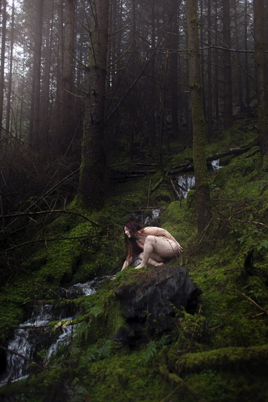 A nude woman crouches on a moss-covered rock in a dense forest near a small stream, surrounded by trees and greenery.