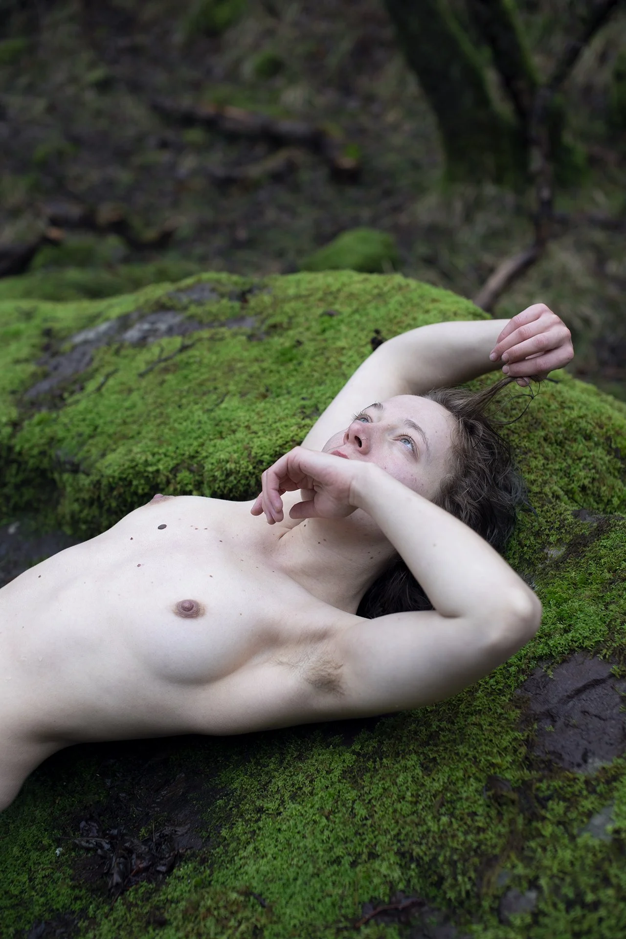 A nude woman with light skin lying on her back on moss-covered ground in a forest, with her arms raised above her head and her eyes looking up.