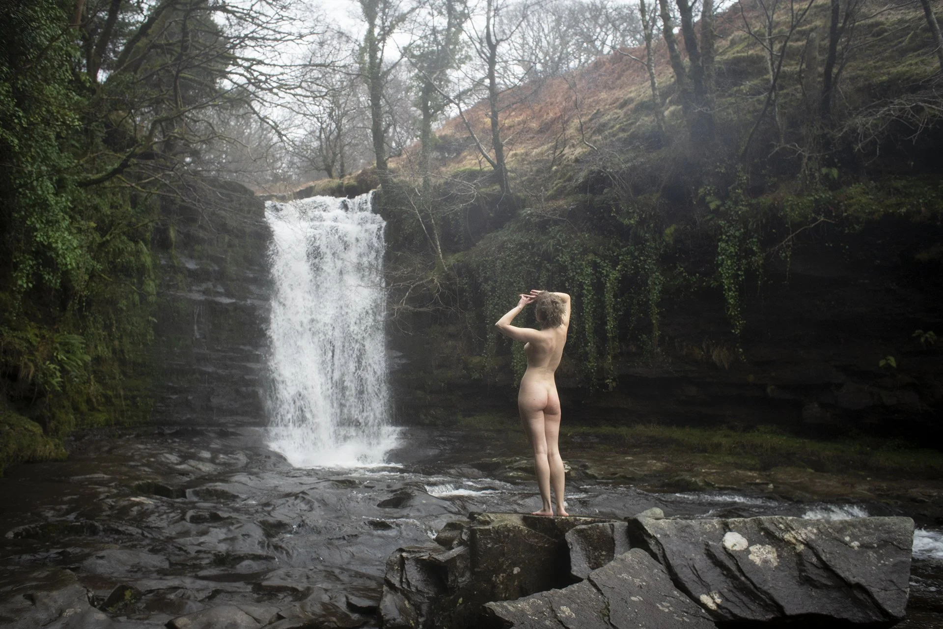 A nude woman standing on rocks near a waterfall in a forested area.