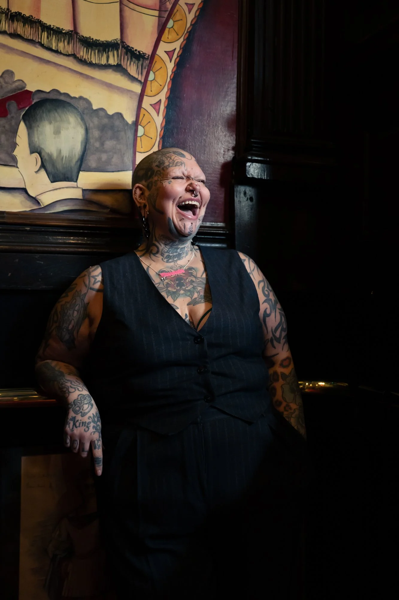 A woman with tattoos and facial piercings sitting indoors, smiling or laughing, with a colorful painting behind her on the wall.