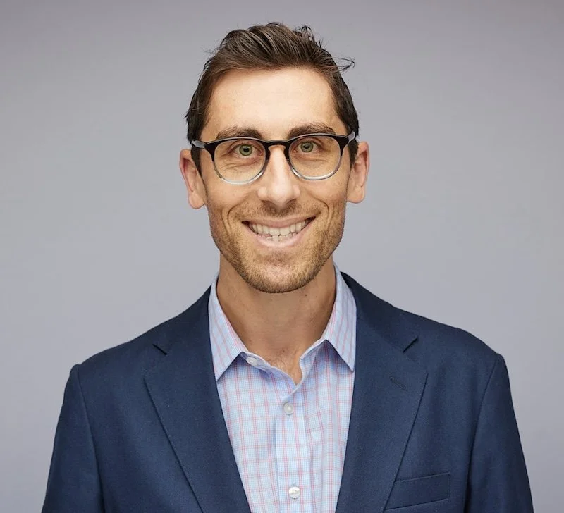 A smiling man in a navy blue suit, light pink checkered shirt, and glasses, against a plain gray background.