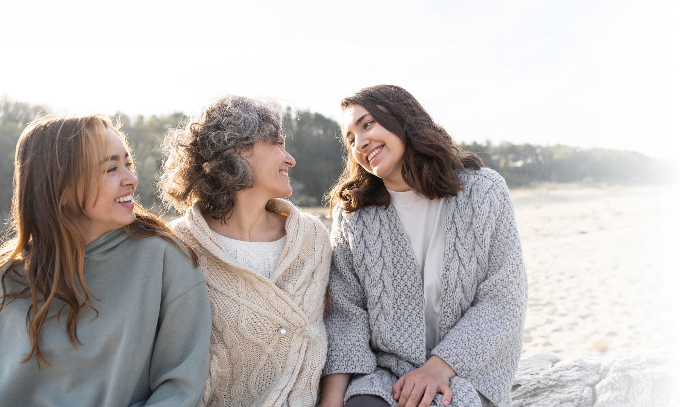 Three women smiling and talking on a beach, with a scenic shoreline and trees in the background.
