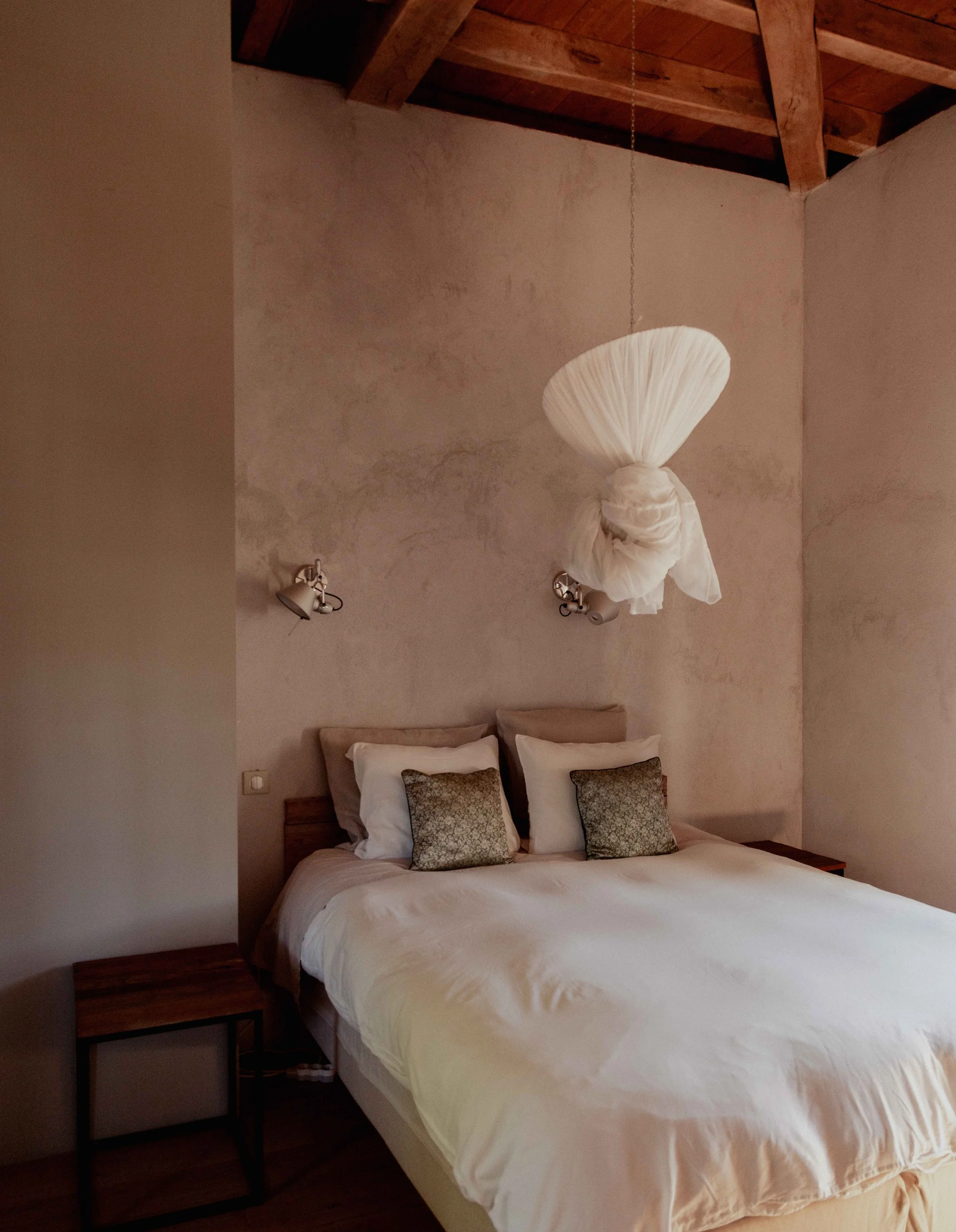 A bedroom with a bed, two pillows, and wall-mounted reading lamps, featuring a unique hanging fabric decoration above the bed.