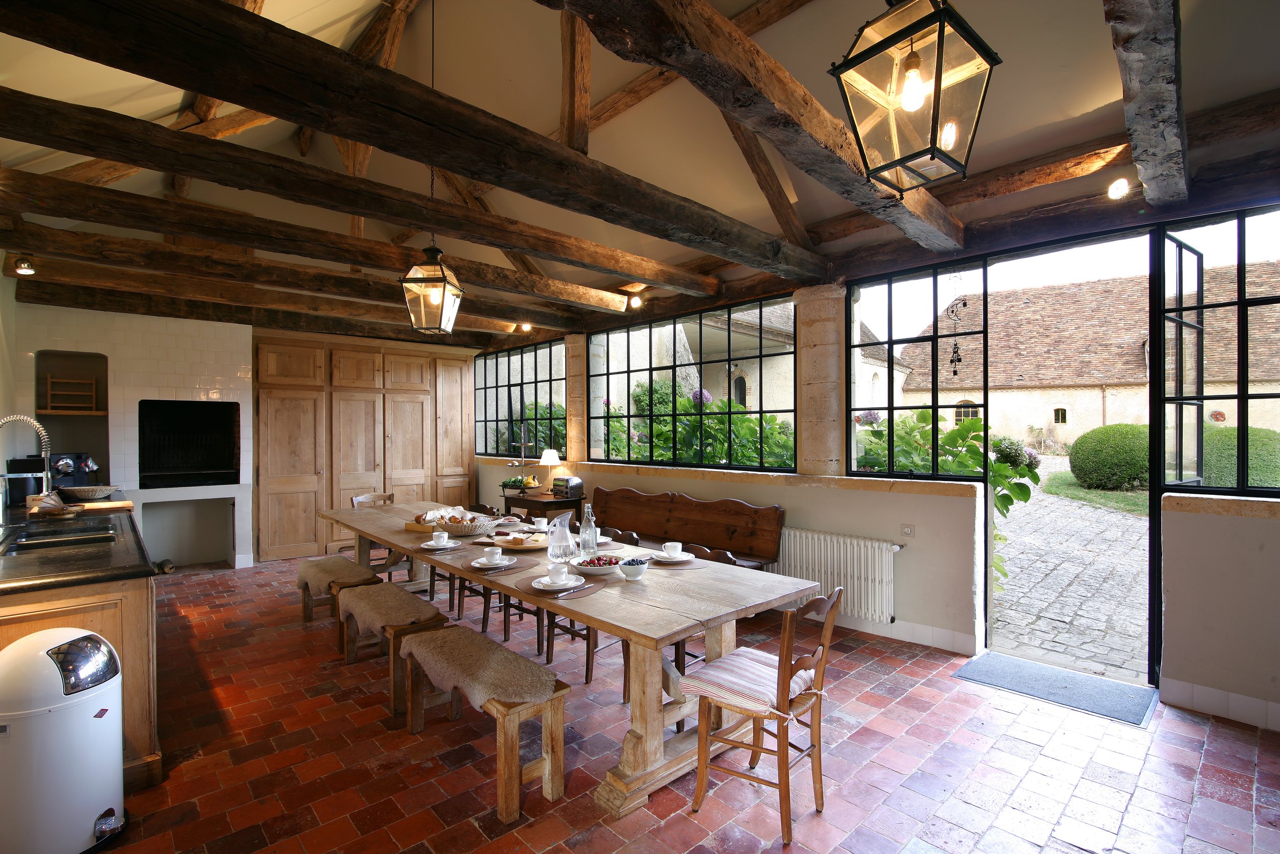 A rustic dining room with exposed wooden beams, large industrial-style windows, a wooden dining table set with dishes and cups, a bench and mix of chairs, and a view of a garden and courtyard outside.