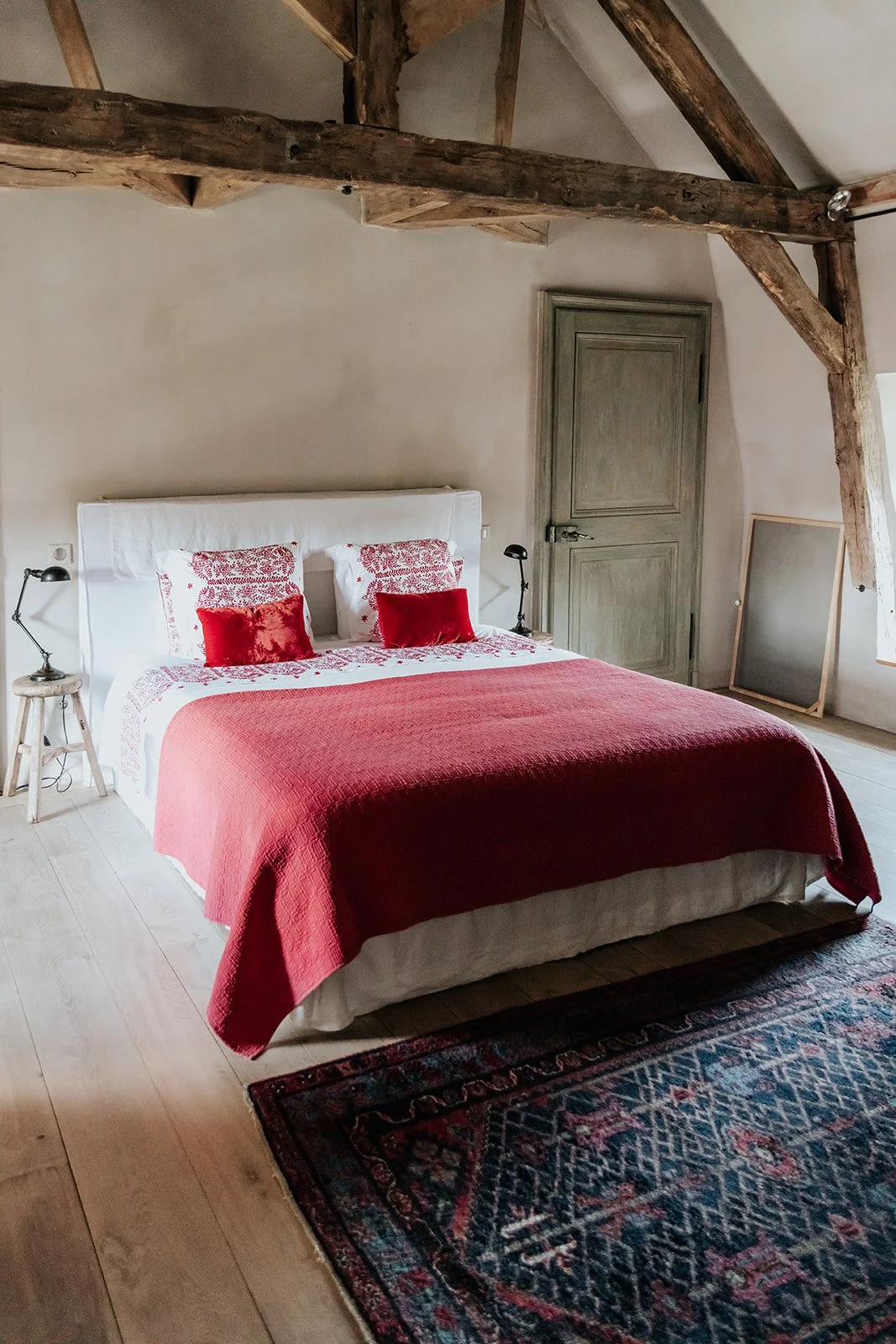A bedroom with a bed, featuring a white headboard, red quilt, and decorative pillows, two bedside lamps, a rustic wooden bench, a mirror, and exposed wooden beams.