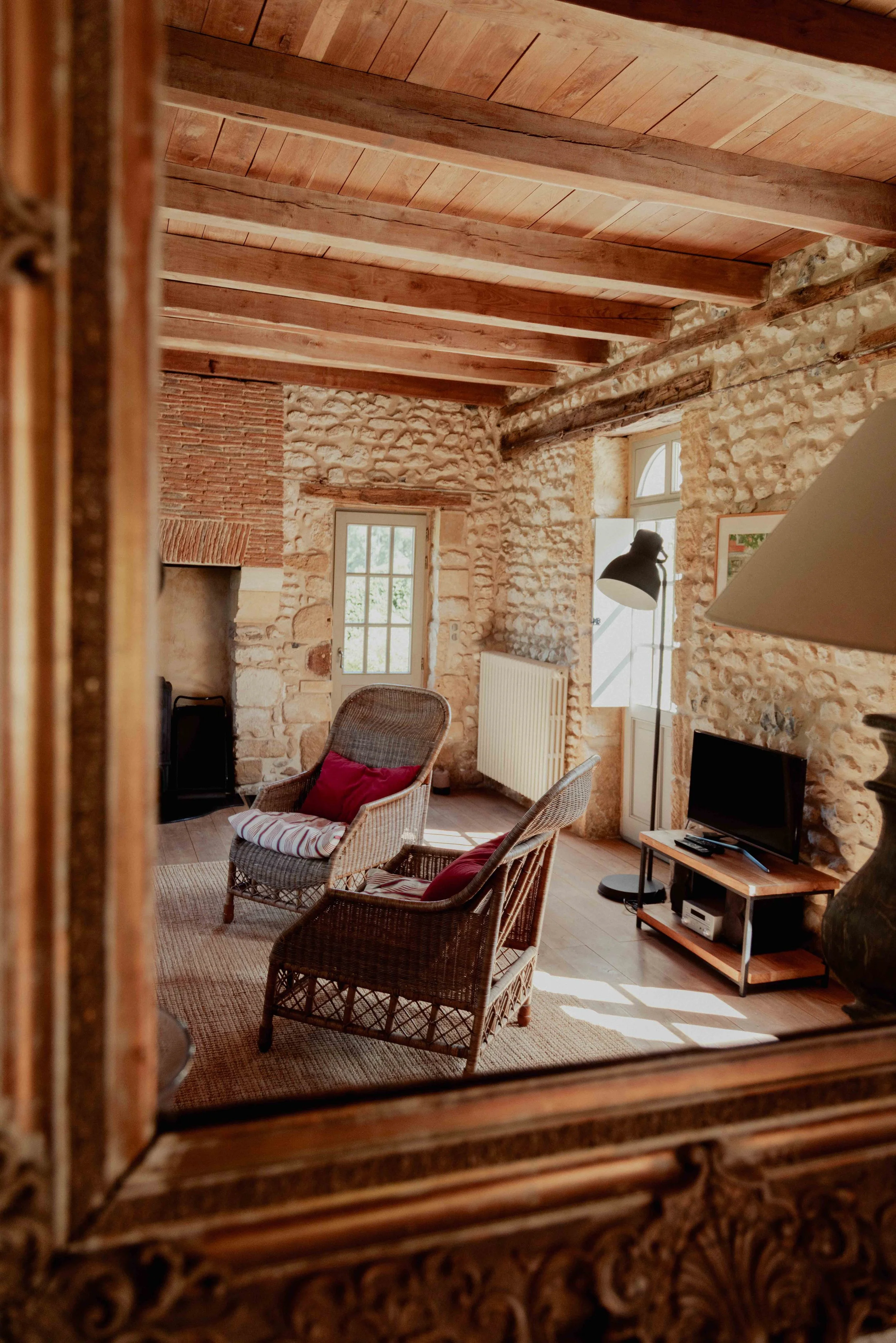 Een woonkamer met een stenen muur, houten plafond, twee wicker stoelen met kussens, een tv op een houten tv-meubel, een staande lamp en een raam dat daglicht binnenlaat.