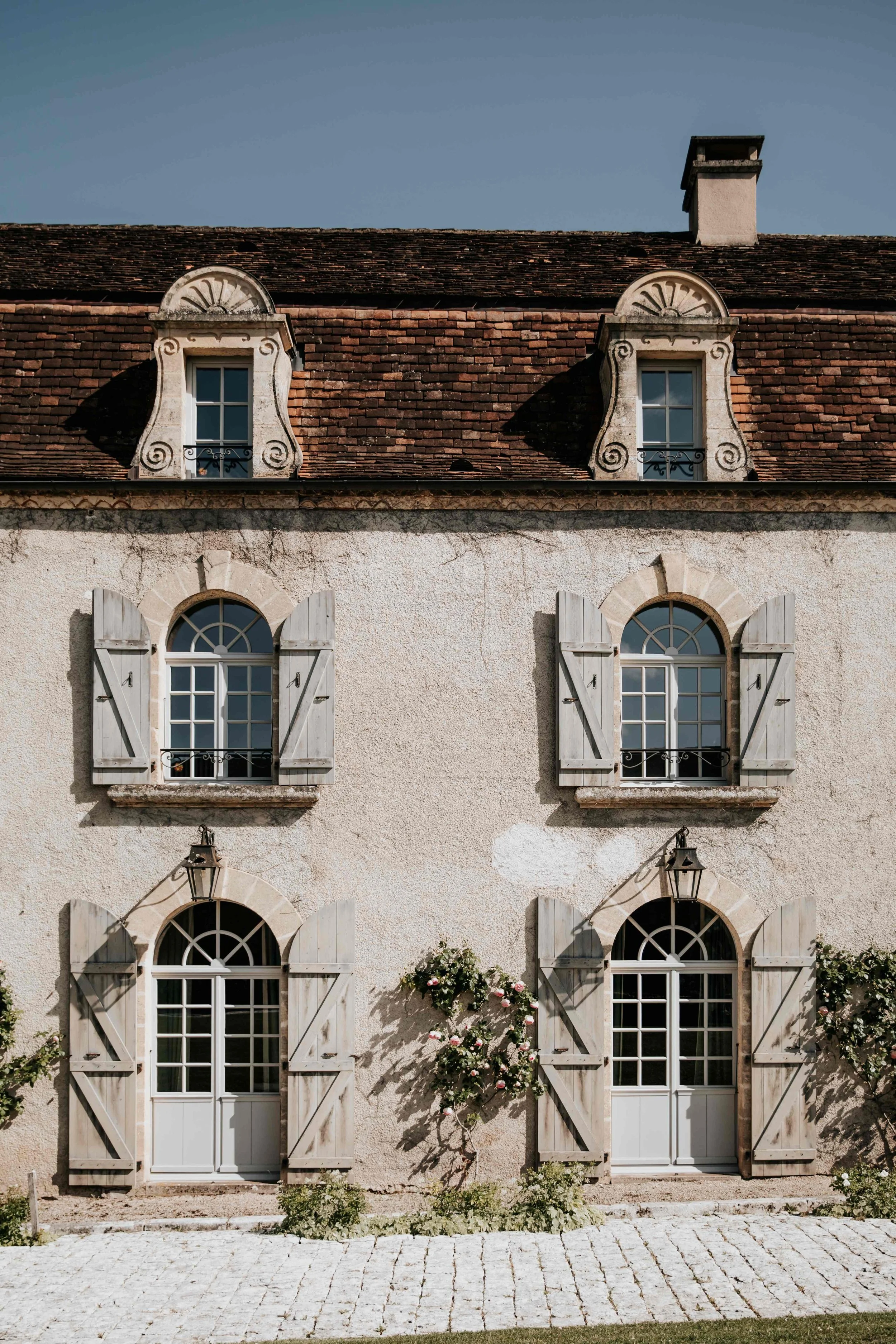 A charming European-style house with a textured stucco exterior, wooden shutters on arched windows, decorative dormer windows on a red-tiled roof, and a small garden with flowering bushes.