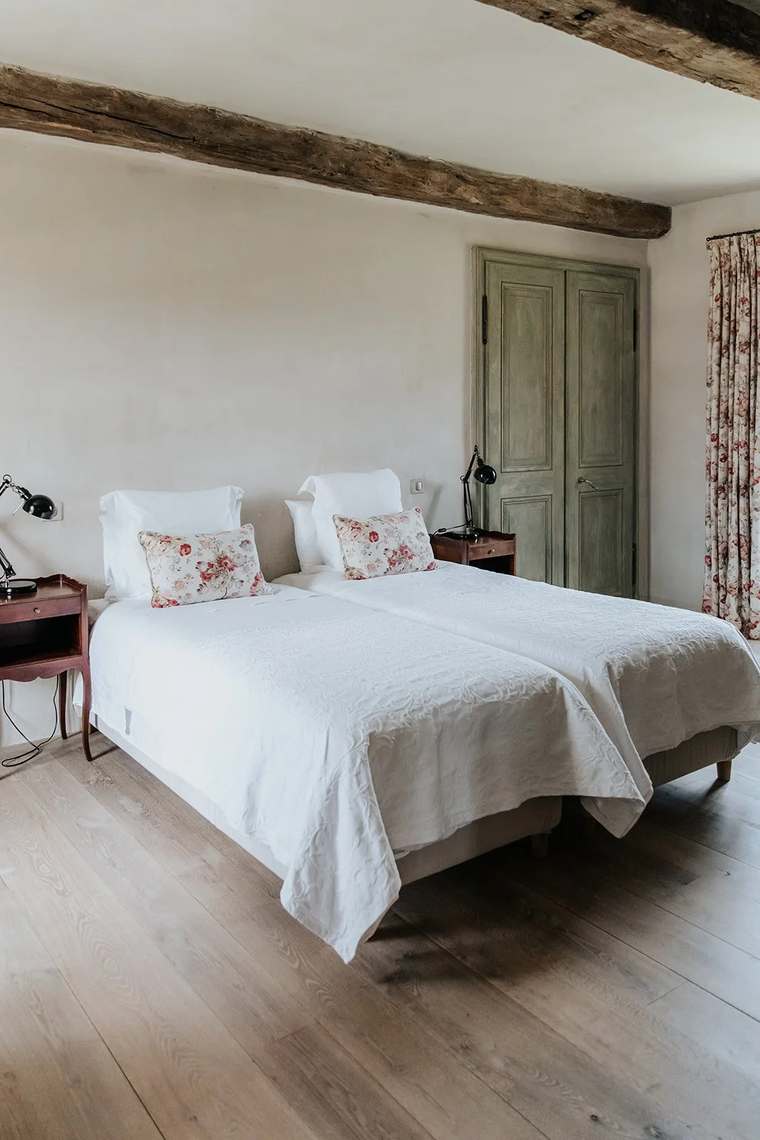 A bedroom with two single beds, each with white bedding, floral pillows, and white headboards. There are two small wooden bedside tables, each with a black lamp. A wooden door and a window with floral curtains are in the background, and the ceiling has exposed wooden beams.