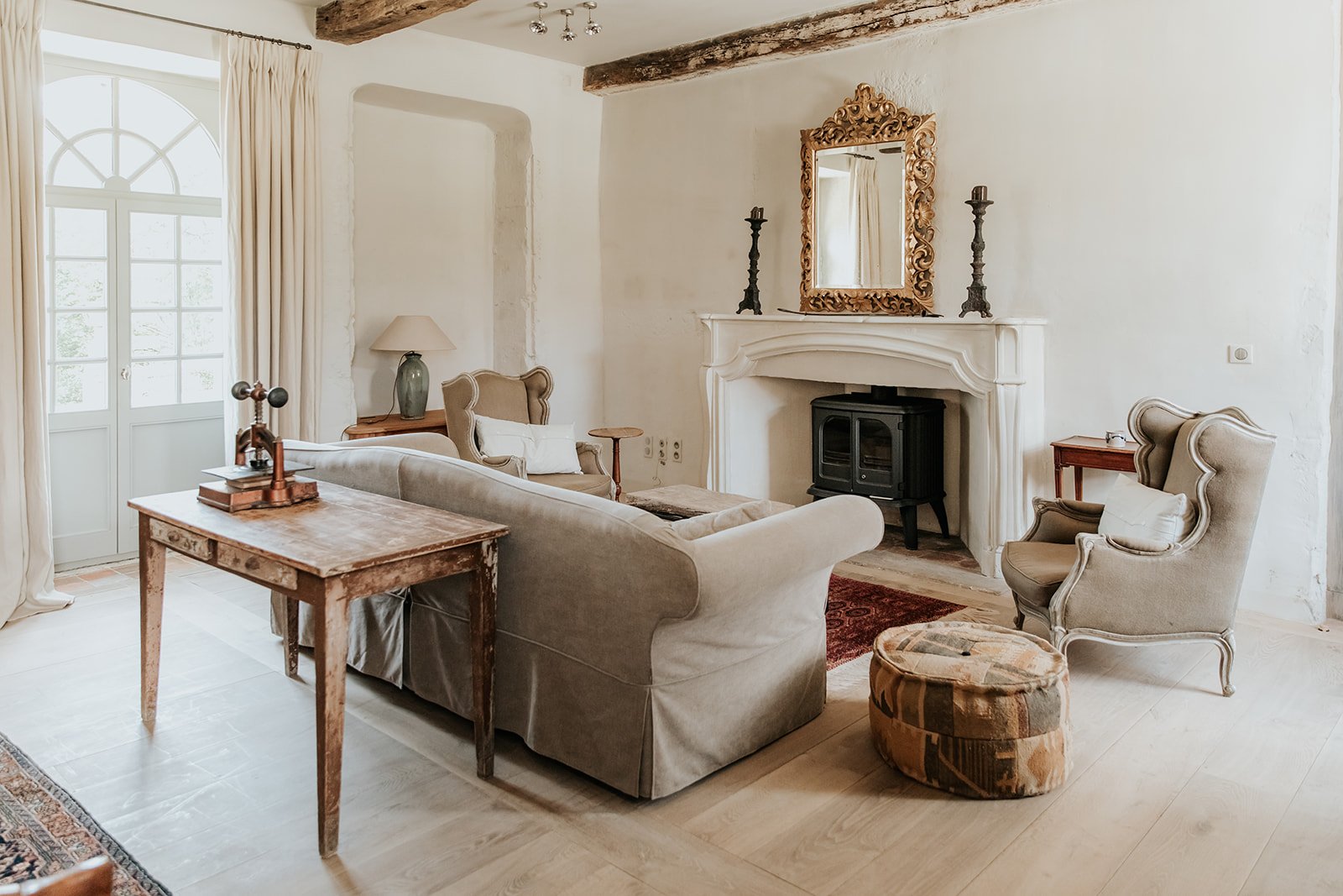Een gezellige woonkamer met een hoge ramen, grote bank, twee elegant gevormde stoel, een deken op een stoel, een rustieke houten salontafel, een muur met een open haard met een spiegel en twee kandelaars erboven, een vloerkleed en decoratieve accessoires.