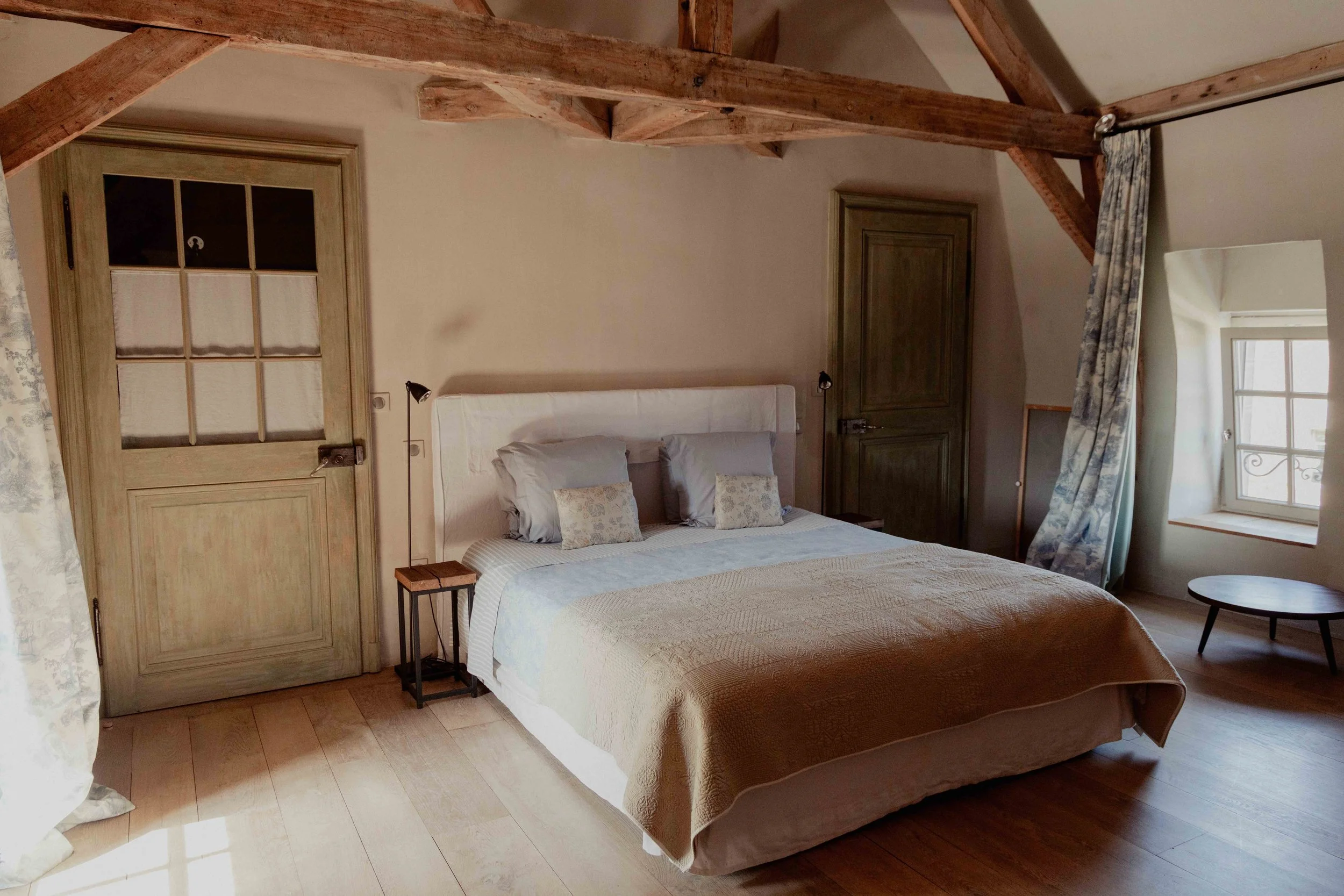 Een slaapkamer met een groot bed, een kaptafel, een nachtkastje, een raam met lichte gordijnen en een deur. Het plafond heeft houten balken.