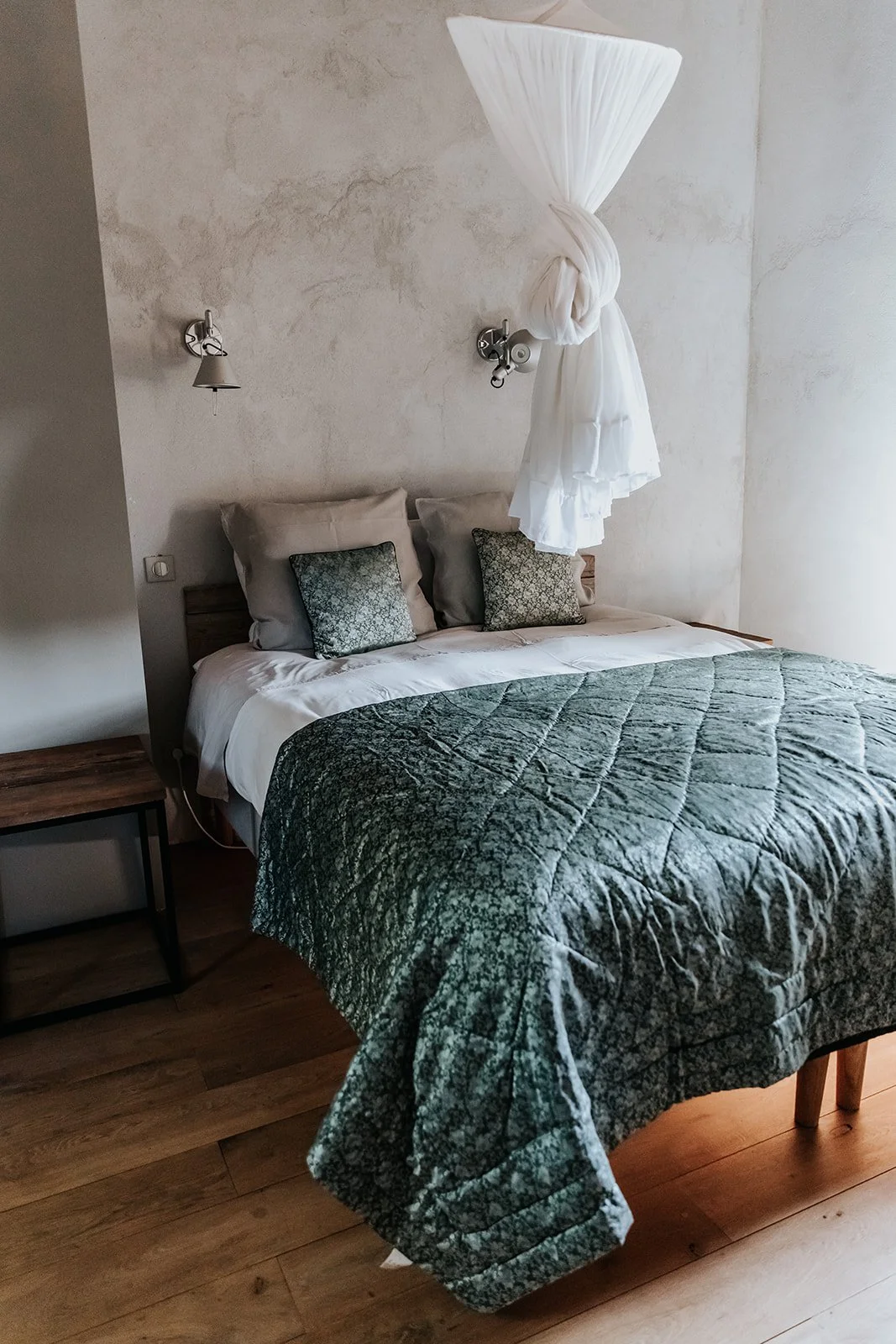 A bedroom with a bed covered in a green bedspread, decorated with pillows, next to a wooden nightstand, with a wall-mounted lamp above the headboard.