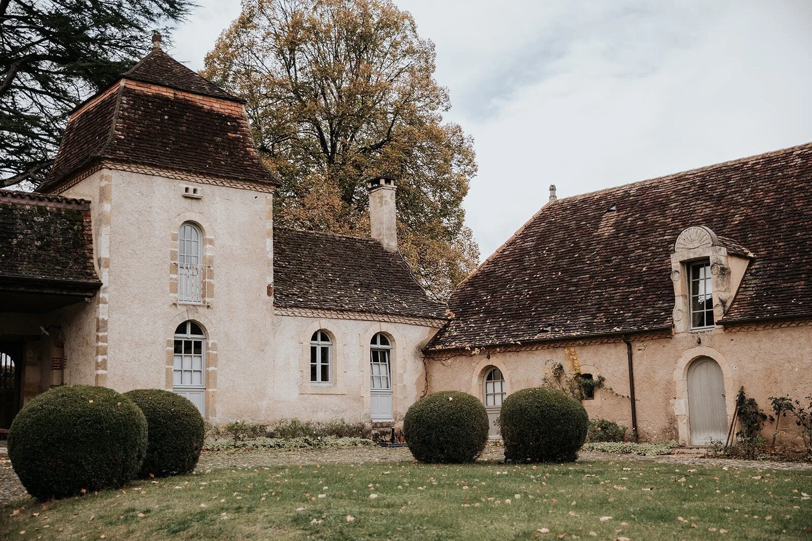 A historic European-style house with a beige stucco exterior, multiple windows, and a steep, brown tile roof. There are four round trimmed bushes in the front yard, and a large tree with golden leaves behind the house. Overcast sky overhead.