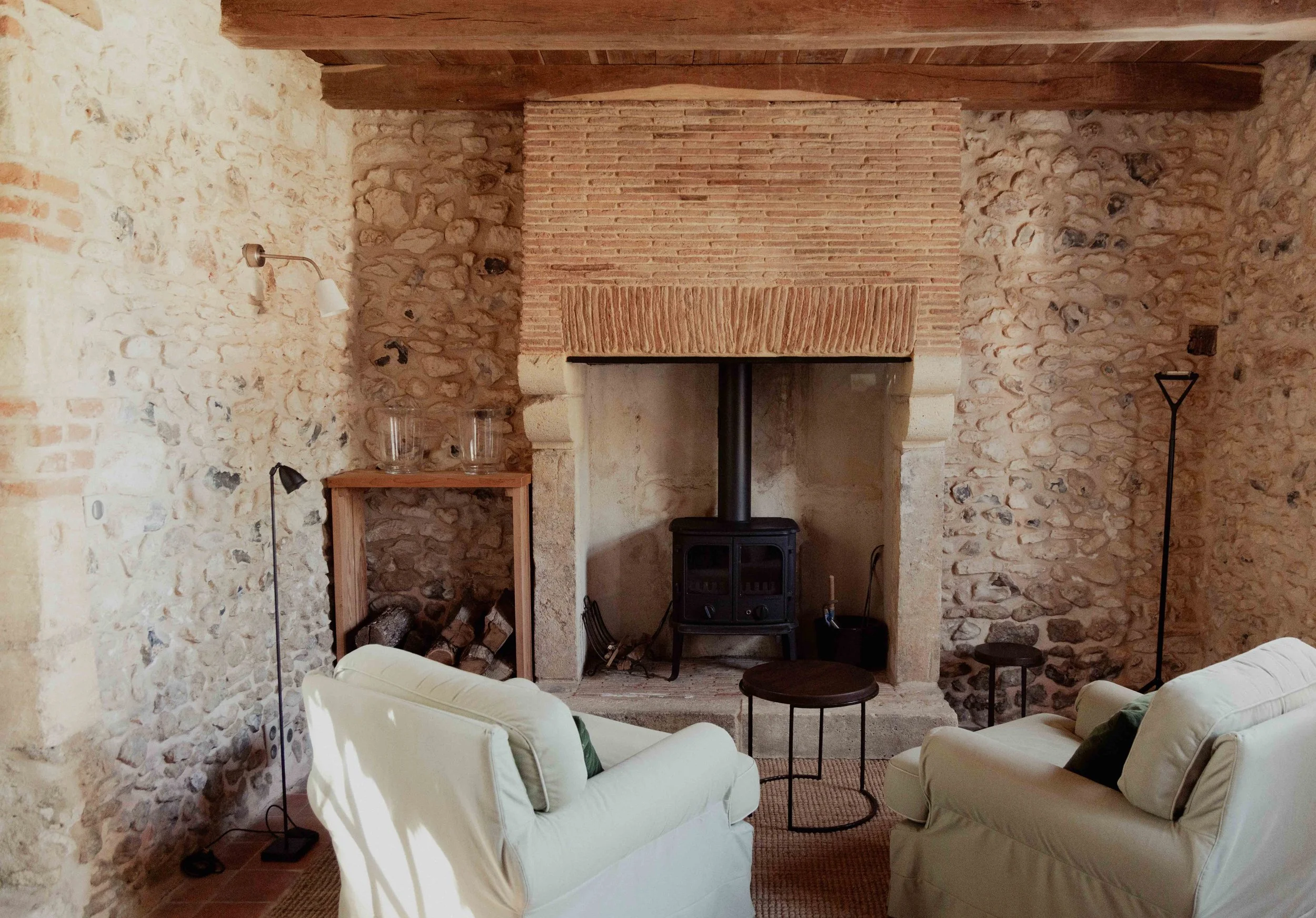 Een gezellige woonkamersetting met een stenen muur en schouw, twee witte fauteuils, een ronde salontafel, een houtkachel, een vloerlamp en een bijzettafel in een rustiek interieur.