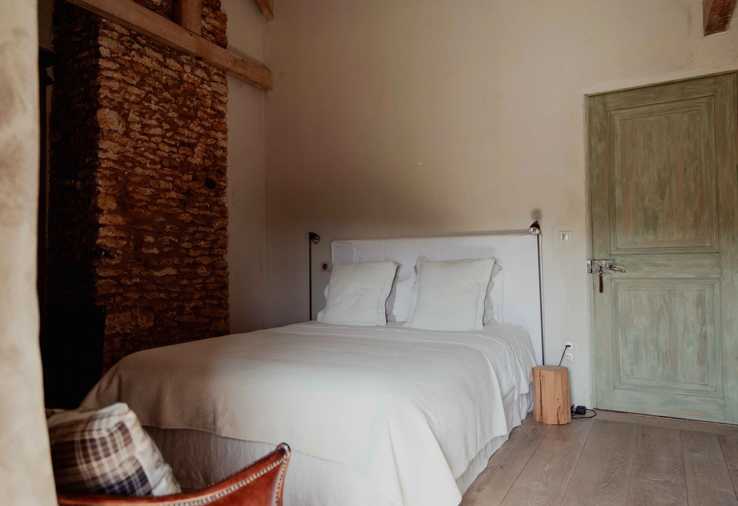 Een slaapkamer met een wit bed, drie witte kussens, een houten nachtkastje, een groene deur, en een muur met een ruwe bakstenen muur en lichte muur.