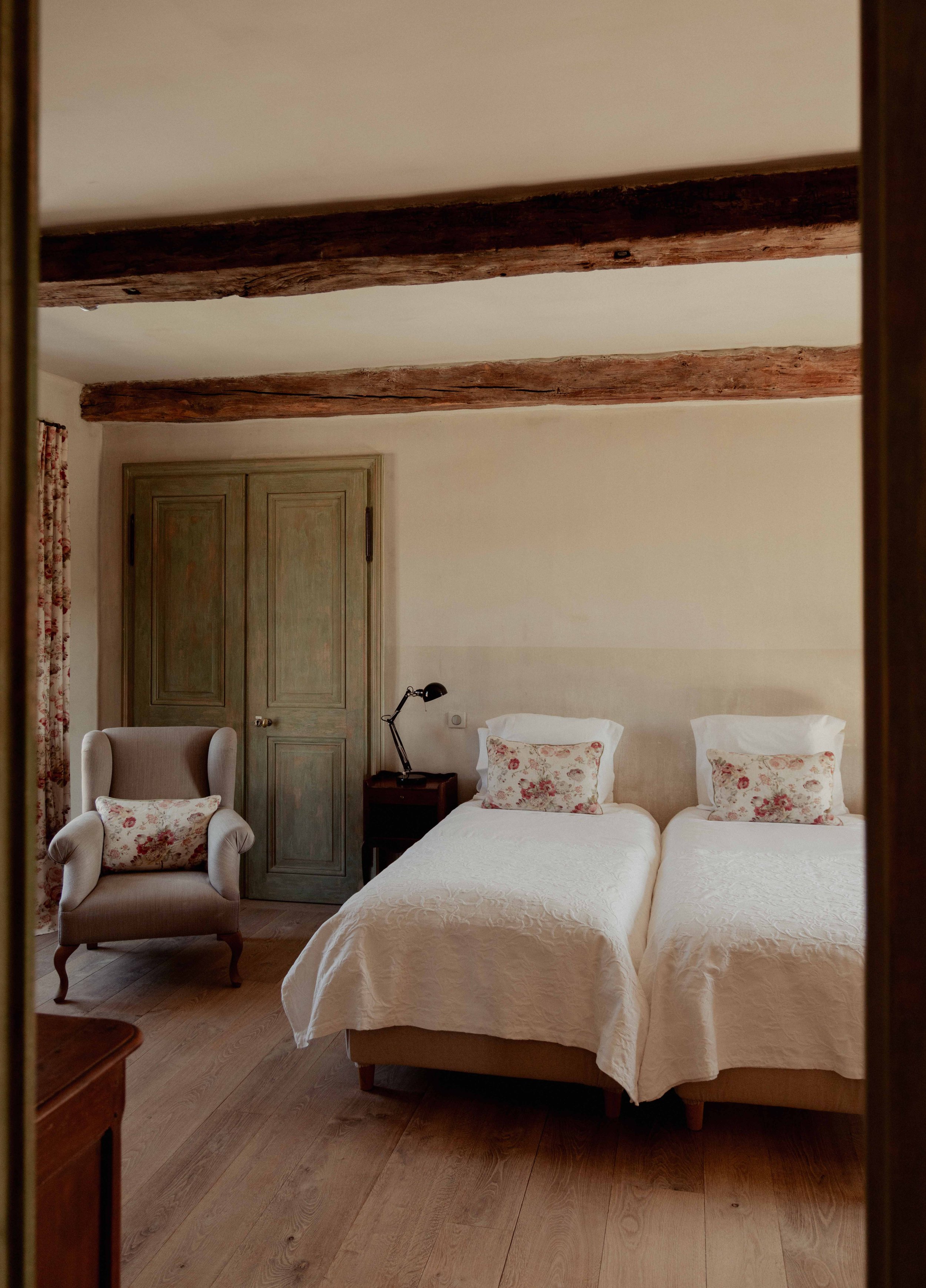 Een slaapkamer met twee eenpersoonsbedden met witte beddengoed en kussens met bloemenpatroon, een beige armstoel met een vergelijkbaar kussen, een nachtkastje met een groene leeslamp, een houten vloer, houten plafondbalken, een groene houten deur en een raam met bloemrijke gordijnen.