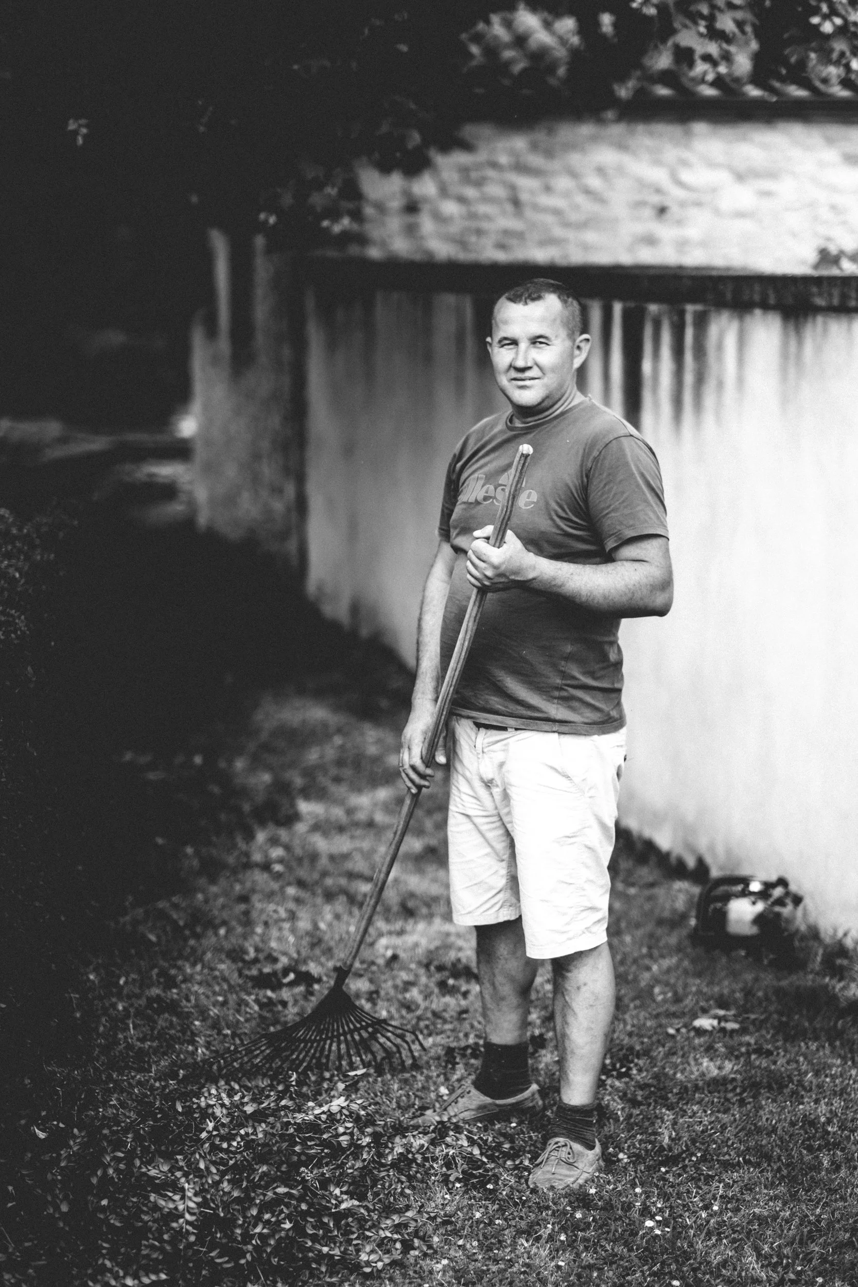 Een man staat buiten, gekleed in een T-shirt, korte broek en sportschoenen, en houdt een grasmaaier of tuingereedschap in zijn rechterhand. Op de achtergrond is een muur en een tuin te zien.
