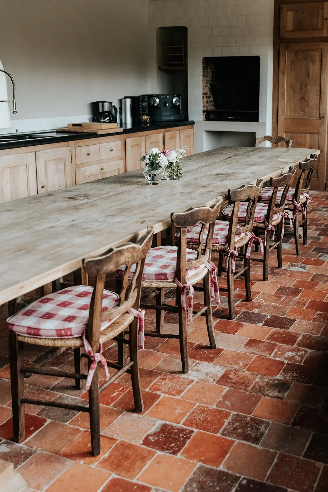 Long wooden dining table with cherry-checked cushioned chairs in a rustic kitchen featuring brick flooring, wooden cabinetry, and a small centerpiece of flowers.
