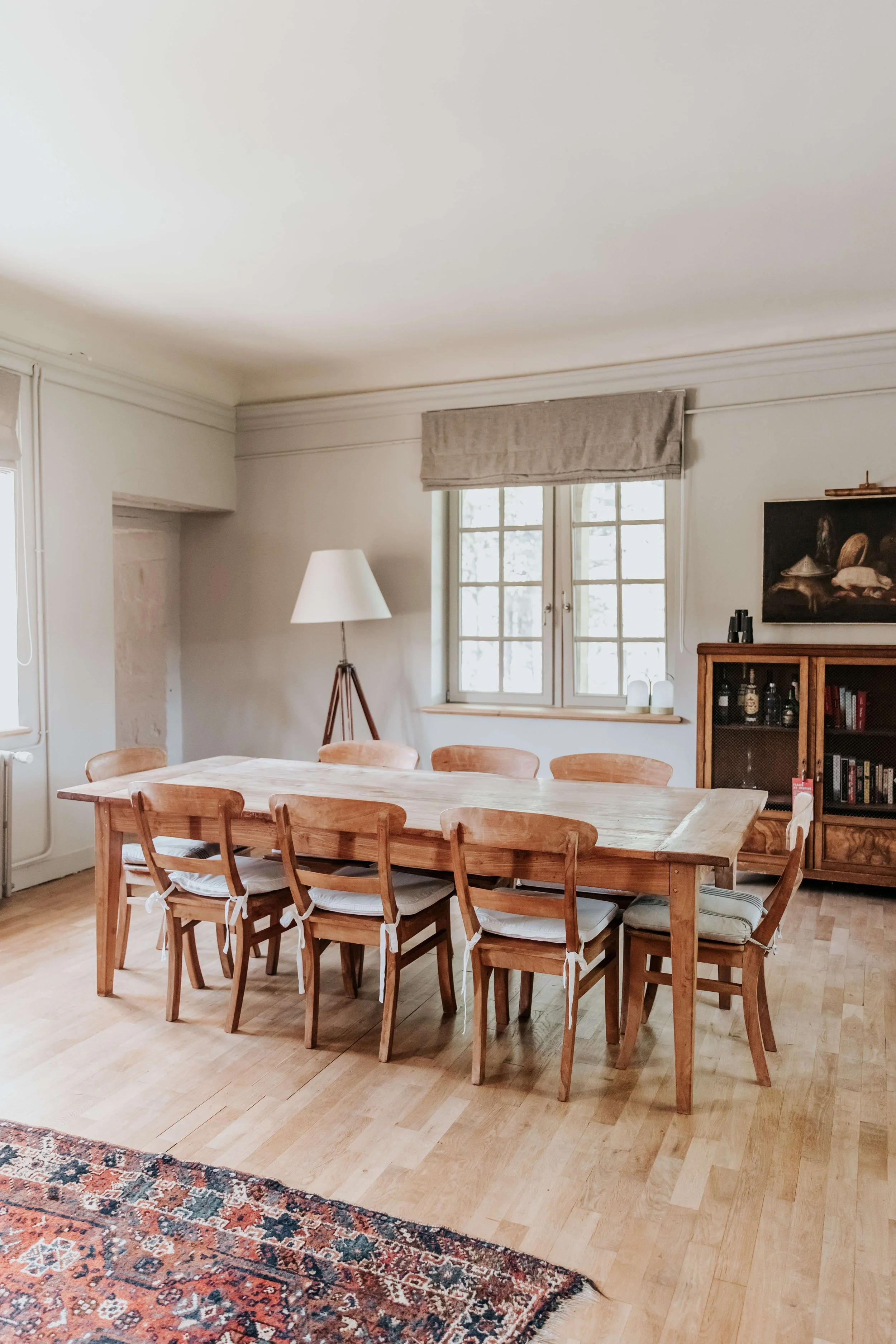 A dining room with a large wooden table, nine wooden chairs with white cushions, a window with a beige Roman shade, a tripod floor lamp, a wooden cabinet with books and bottles, and a partially visible patterned rug.