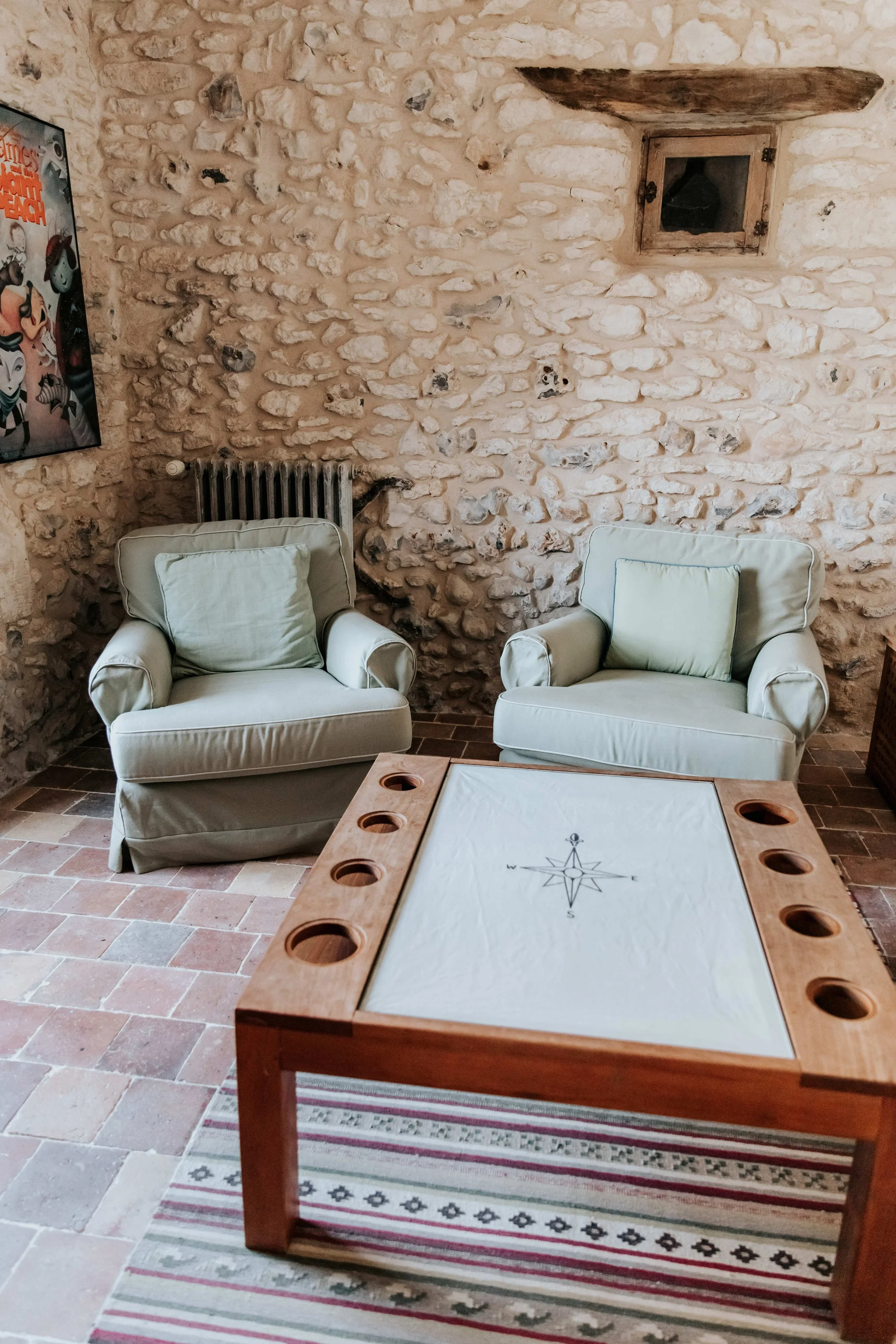 A cozy sitting area with two light green armchairs, a wooden table with cup holders, stone wall with a small wooden window, and a colorful patterned rug.