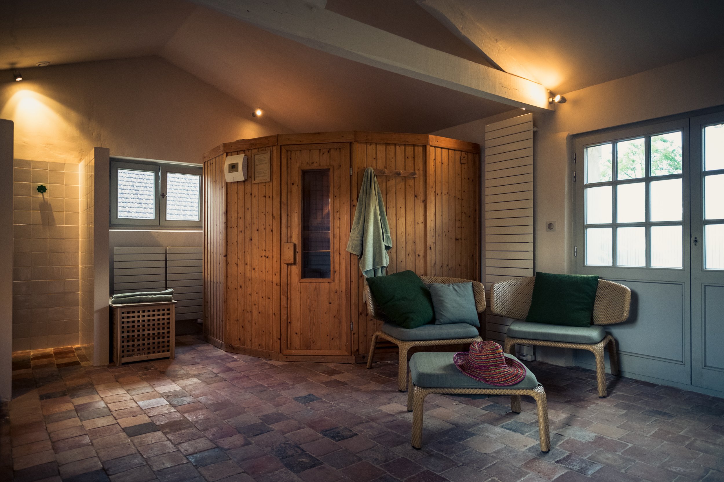 Interieur van een wellnessruimte met een houten sauna, twee witte fauteuils met kussens, een voetenbankje met een kleurrijke hoed, en grote ramen met uitzicht op groen.