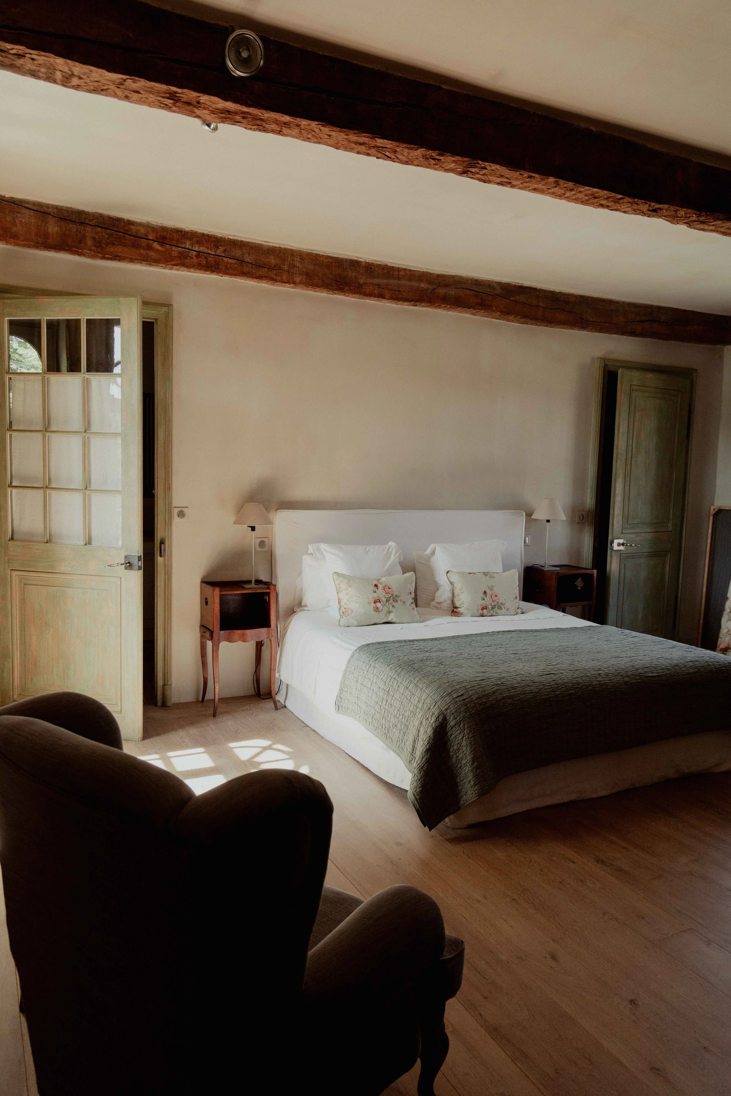 A cozy bedroom with a large bed, decorated with white pillows and floral pillows, a gray blanket, and wooden nightstands with lamps on each side. There is a green armchair in the foreground, and wooden beams on the ceiling with natural light coming through a partially open door.
