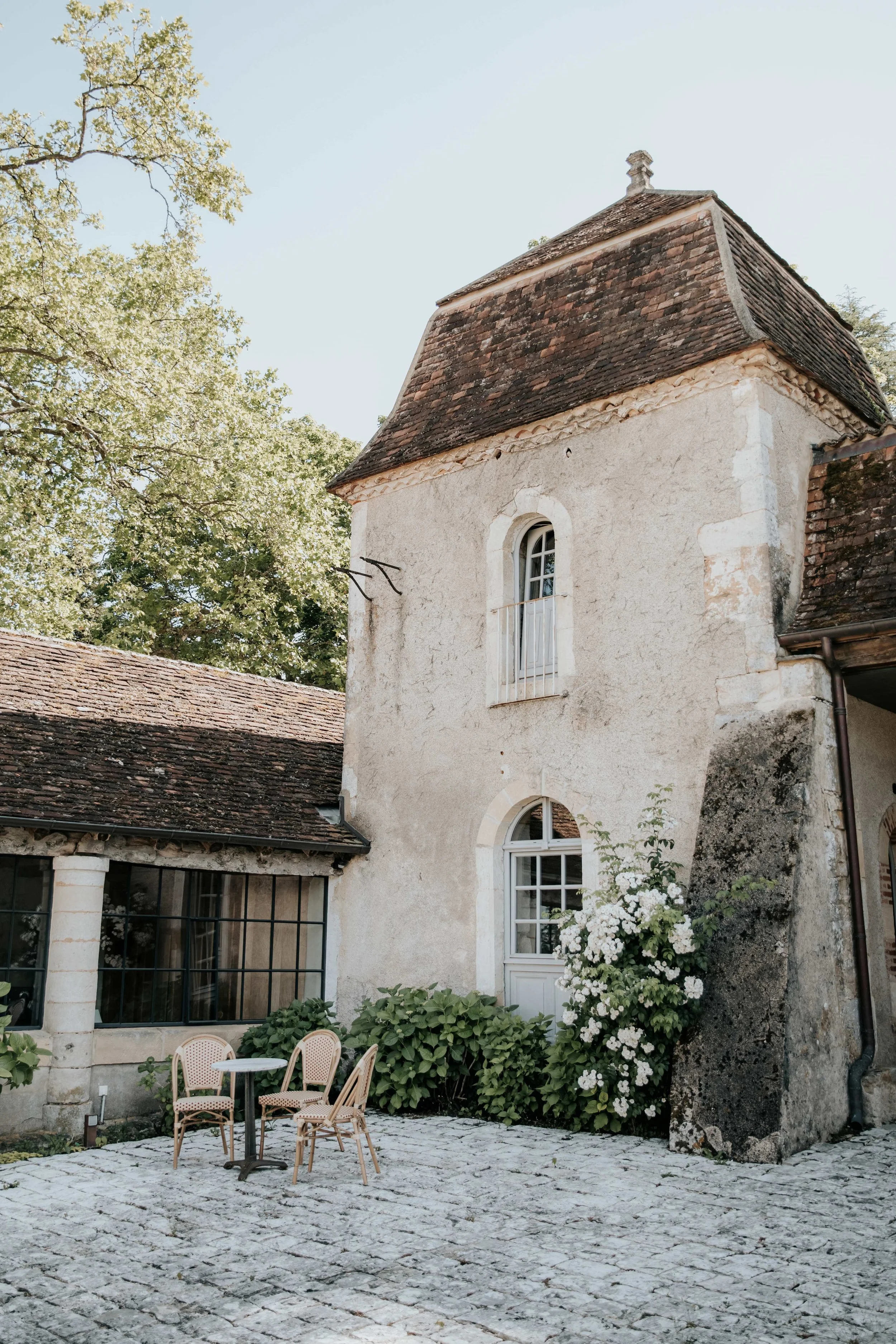 An old European building with white stucco walls, arched windows, and a red tiled roof, surrounded by greenery and white flowering bushes, with outdoor seating on a cobblestone patio.