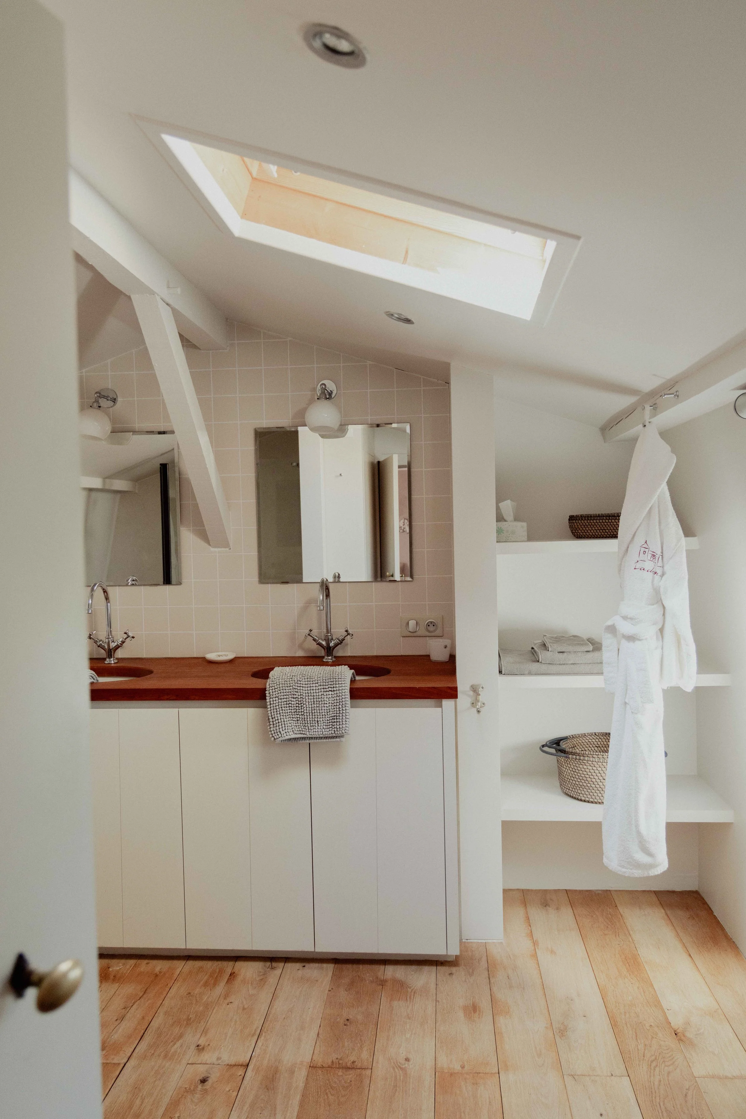 Badkamer met dubbele wastafel, spiegel, en witte kasten onder de wasbakken. Een raam in het plafond voor natuurlijk licht. Een badjas hangt aan een rail, en er zijn open planken met handdoeken en manden.