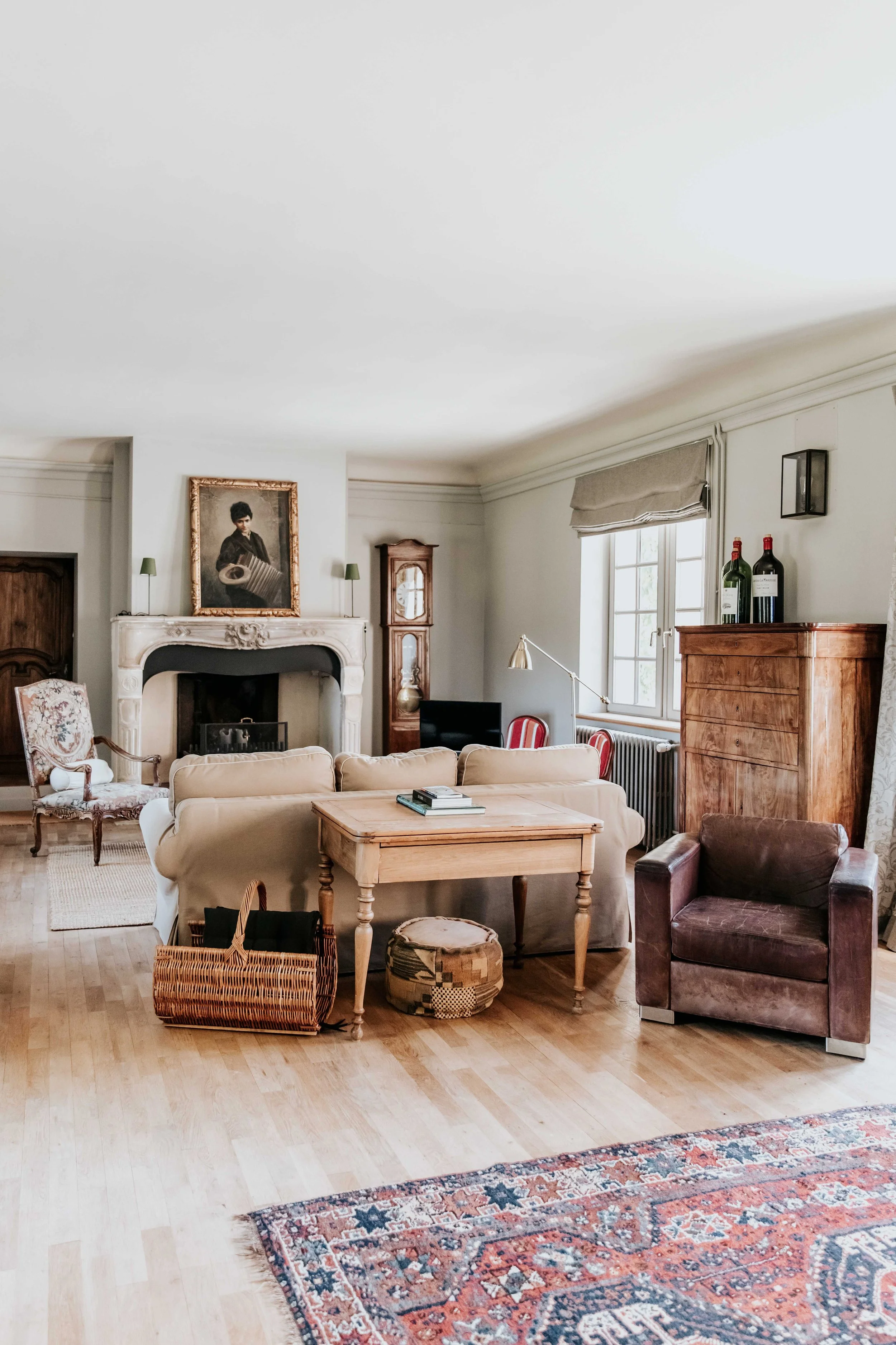 A cozy living room with a beige sofa, a dark armchair, a wooden sideboard, a vintage armchair, a fireplace with a portrait above it, a wooden armchair, a wicker basket, a round ottoman, a wooden coffee table, and a patterned rug. There are two bottles of wine and a window with a beige Roman shade.