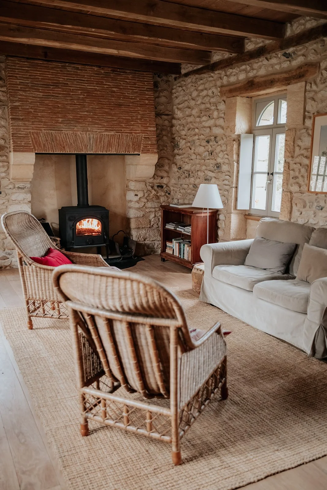 Warme woonkamer met stenen muren, houten plafond en een haard met een brandend vuur, ingericht met wicker stoelen, een witte bank, een houten boekenrek en een witte tafel lamp.