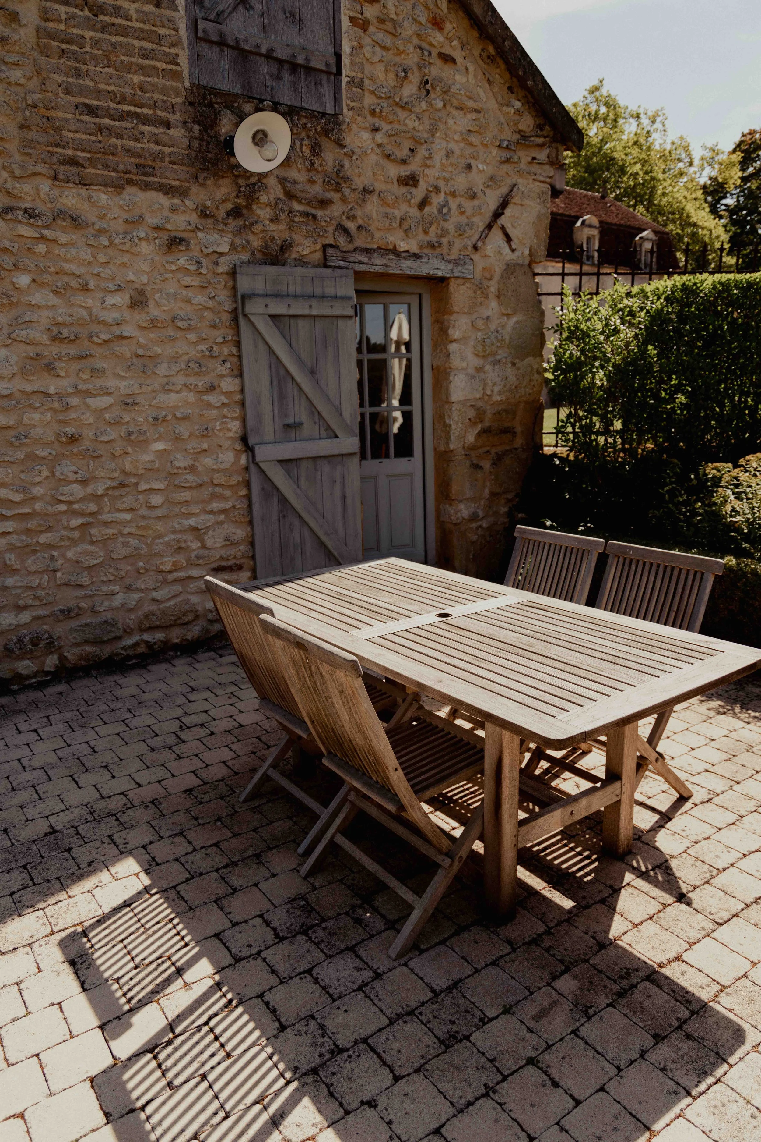 Een houten tuinset met tafel en vier stoelen op een stenen terras naast een huis met een stenen muur en houten luikjes op de ramen.