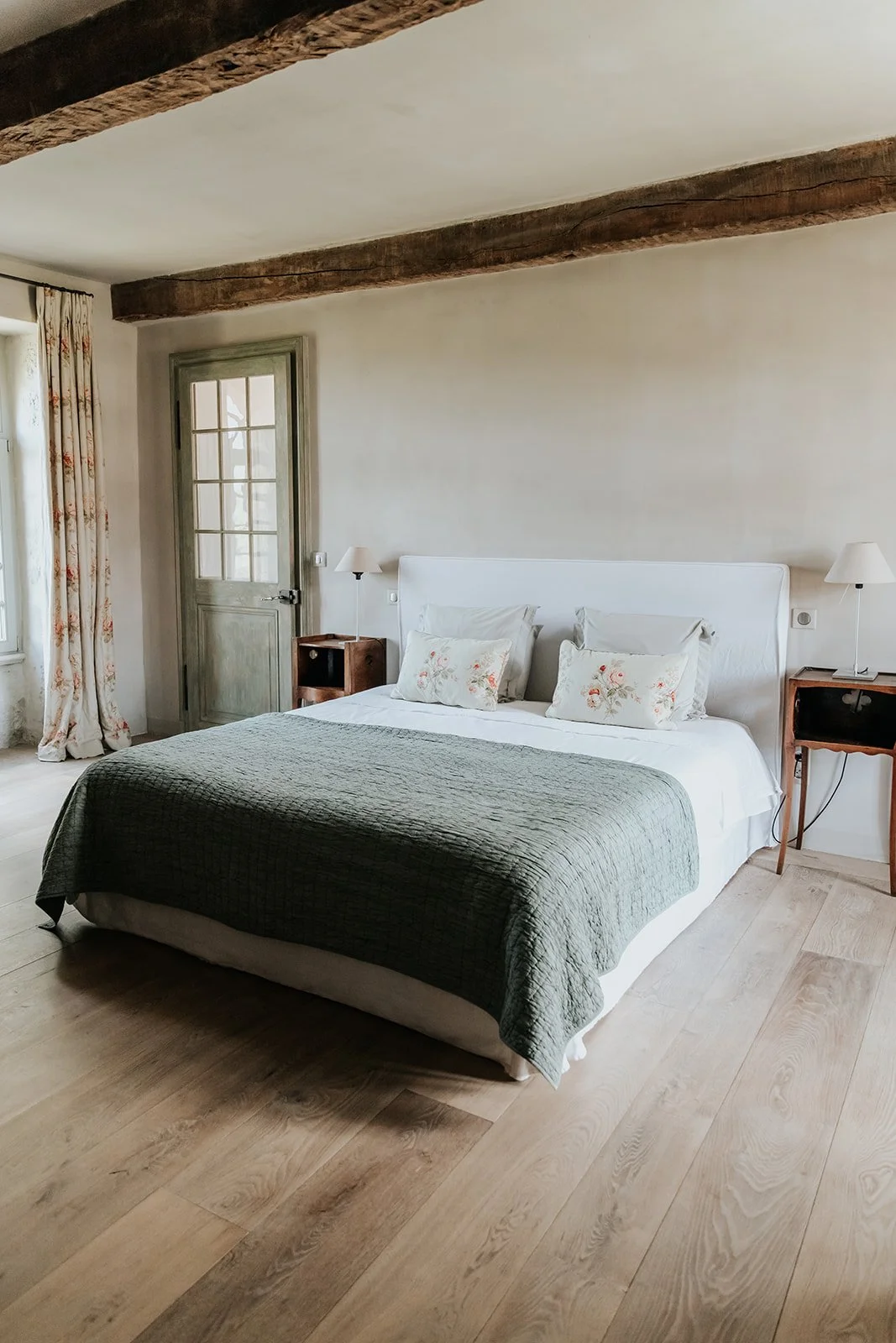 A cozy bedroom with a large bed covered with a green quilt, white pillows, and floral decorative pillows. There are two nightstands with lamps on either side of the bed. The room features wooden beams on the ceiling, a window with floral curtains, and a door with glass panels, all with a rustic style.