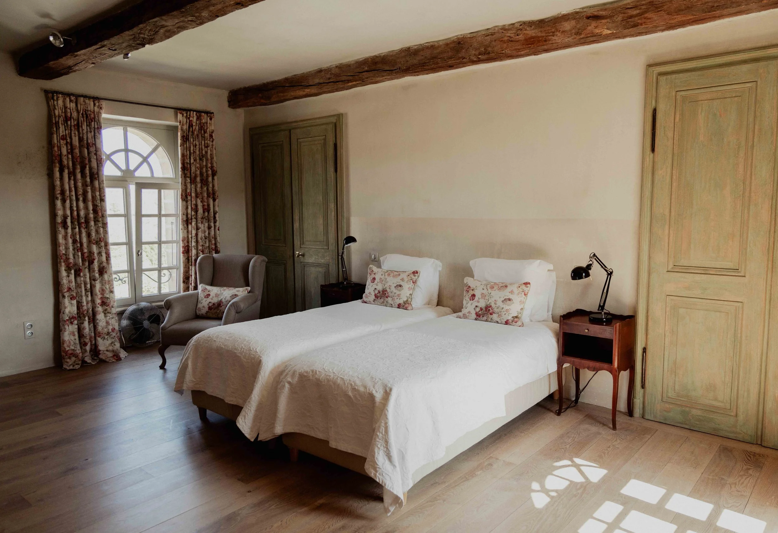 Slaapkamer met twee bedden, een grote venster met bloemen gordijnen, een fauteuil met kussen, twee nachtkasten met leeslampen, houten vloeren, oude houten balken op het plafond, een houten deur en een ventilator.