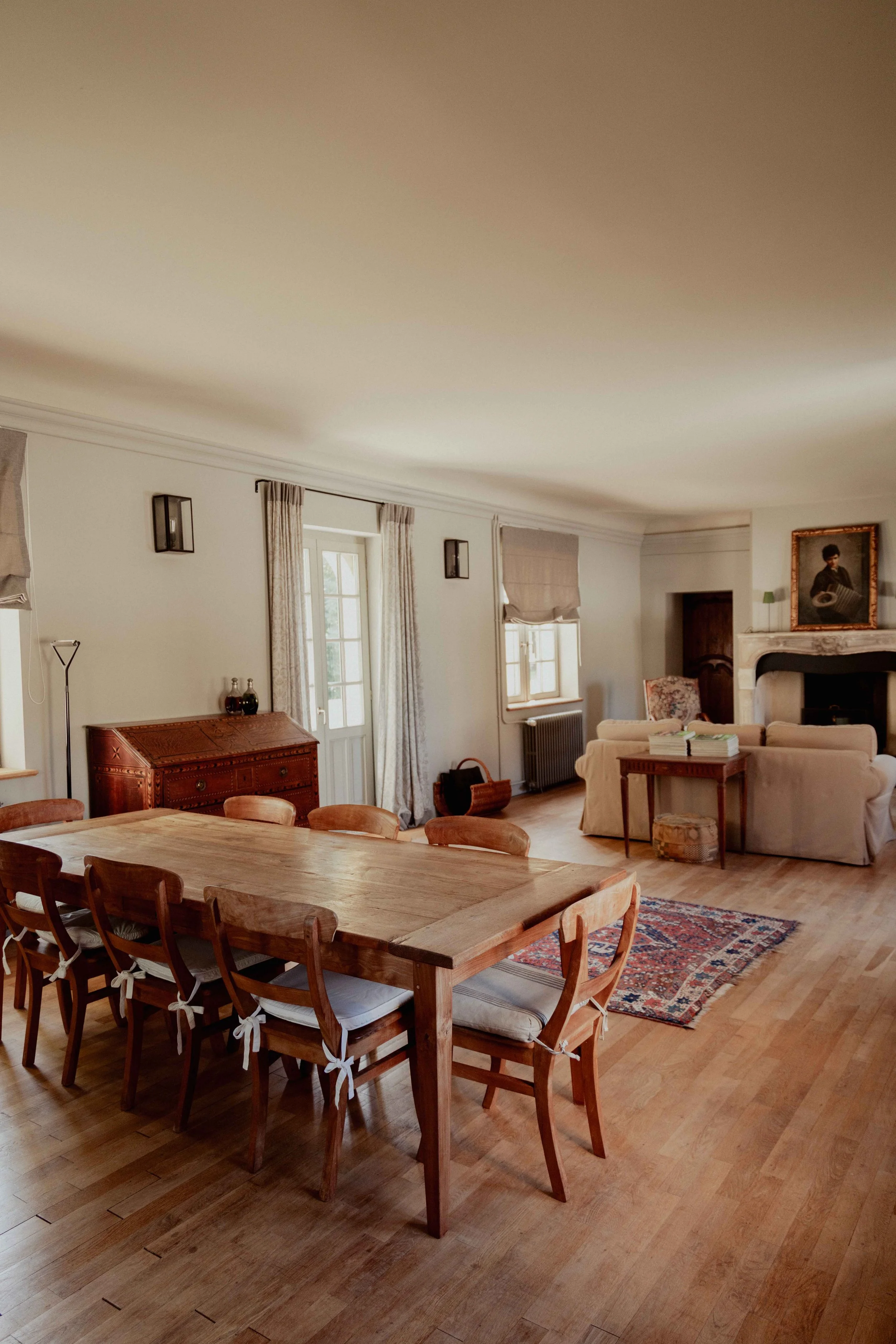 Een ruim woonkamer met een houten eettafel en leren stoelen, een zithoek met banken en een open haard, ramen met gordijnen en een schilderij boven de open haard.