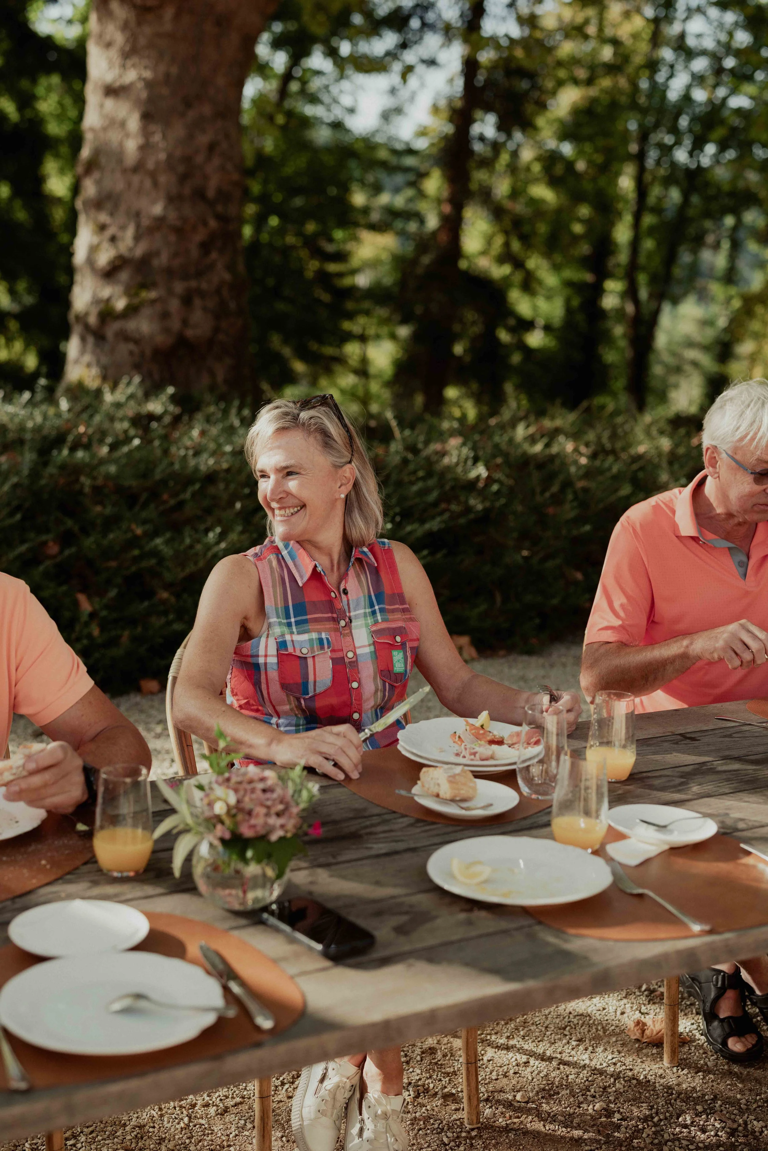 A group of older adults sitting at an outdoor wooden dining table with plates, glasses, and a flower arrangement, enjoying a meal in a wooded area.