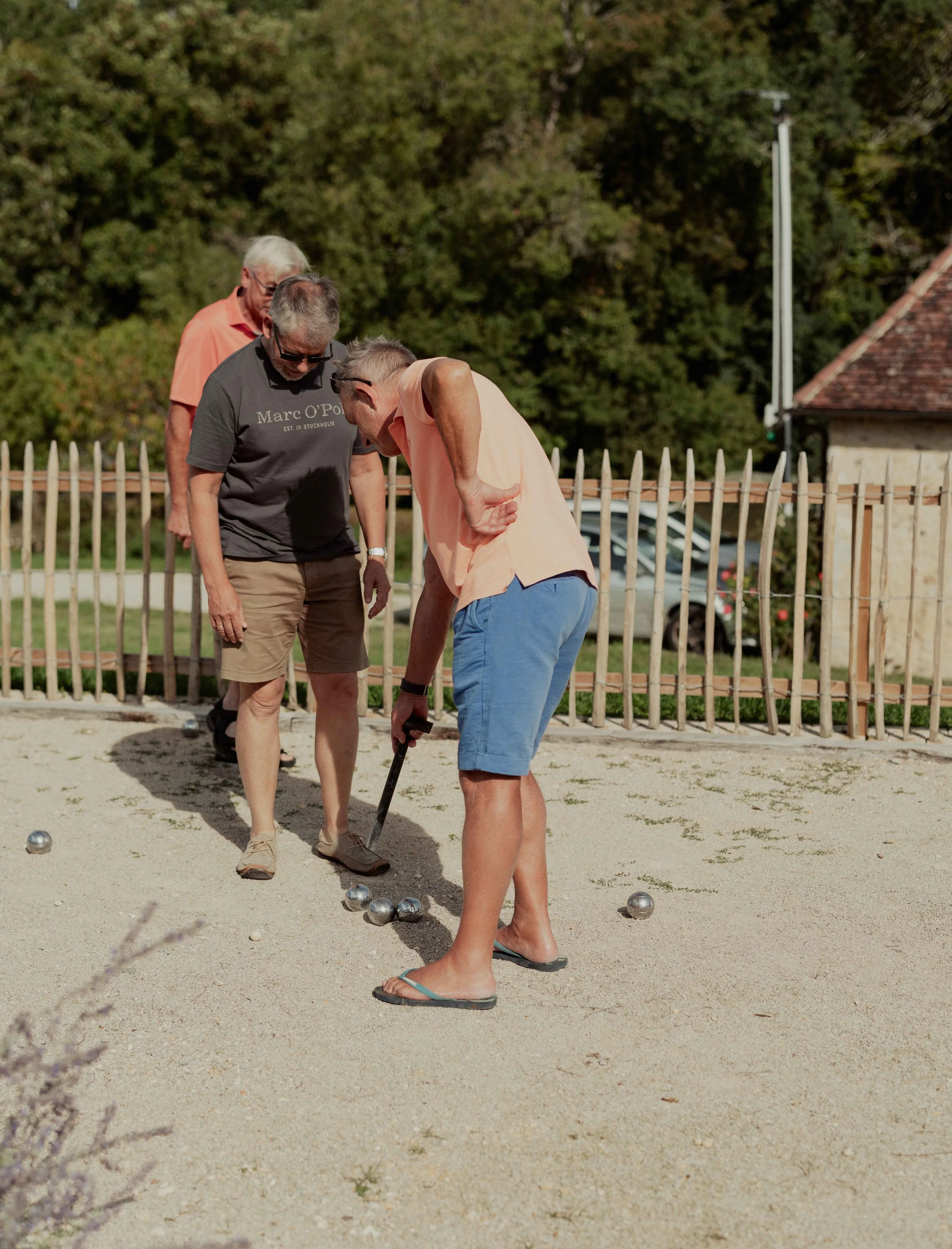 Three older men playing pétanque on a sandy court, with a wooden fence and trees in the background.