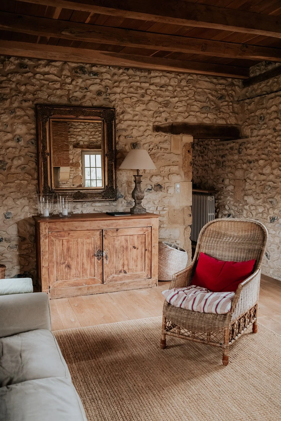 Interieur van een gezellige kamer met een stenen muur, houten plafond, een houten kast met een lamp en een spiegel, een wicker fauteuil met een rood kussen, een beige bank en een wollen vloerkleed.