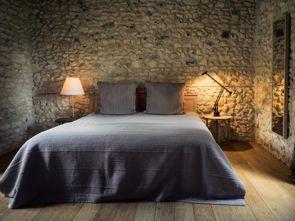 Een slaapkamer met een bed, twee sierkussens, een nachtkastje met een lamp en een verstelbare bureaulamp, met een stenen muur en houten vloer.