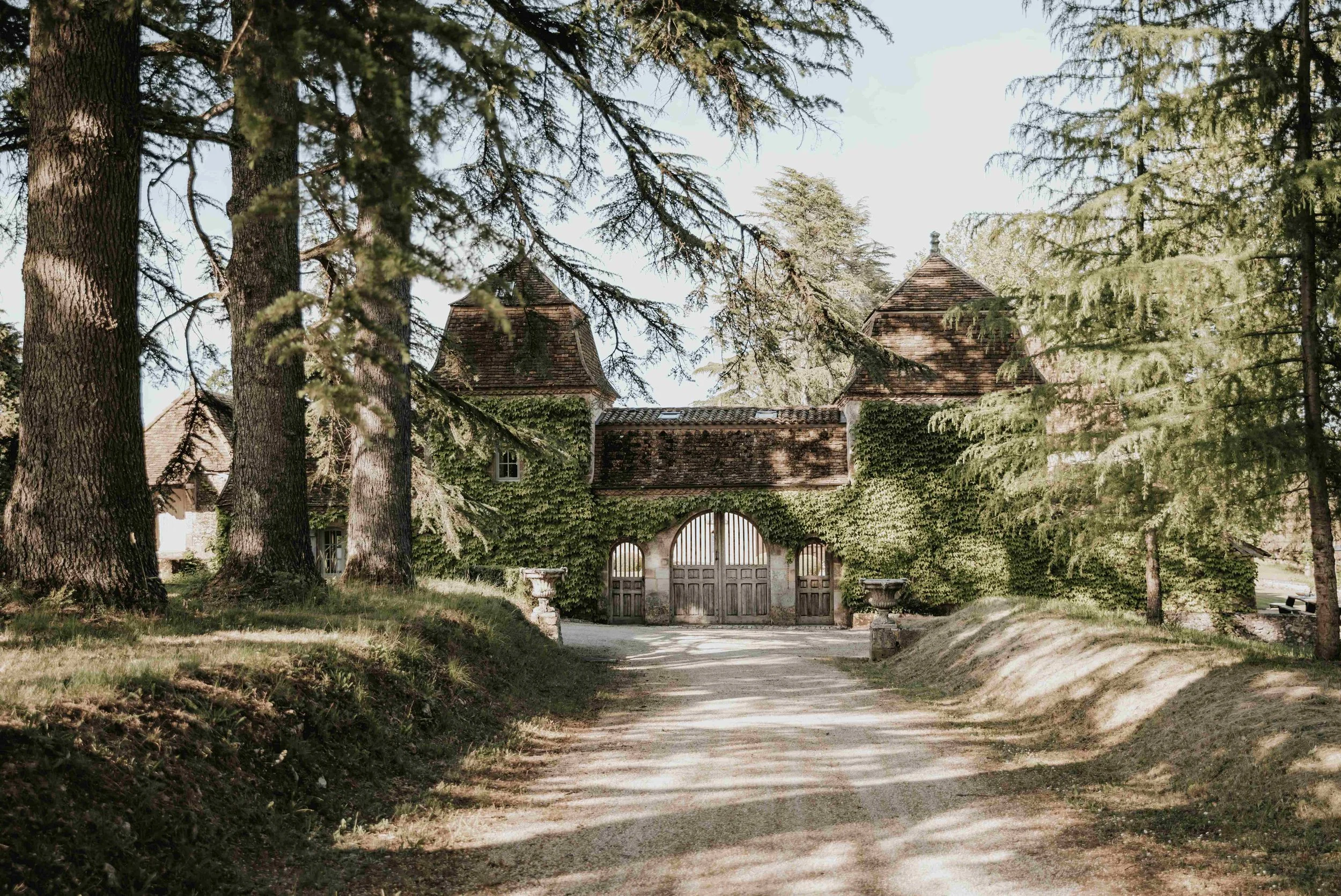 A dirt driveway leading up to an old stone gatehouse covered in green ivy, surrounded by tall trees with sunlight filtering through the branches.