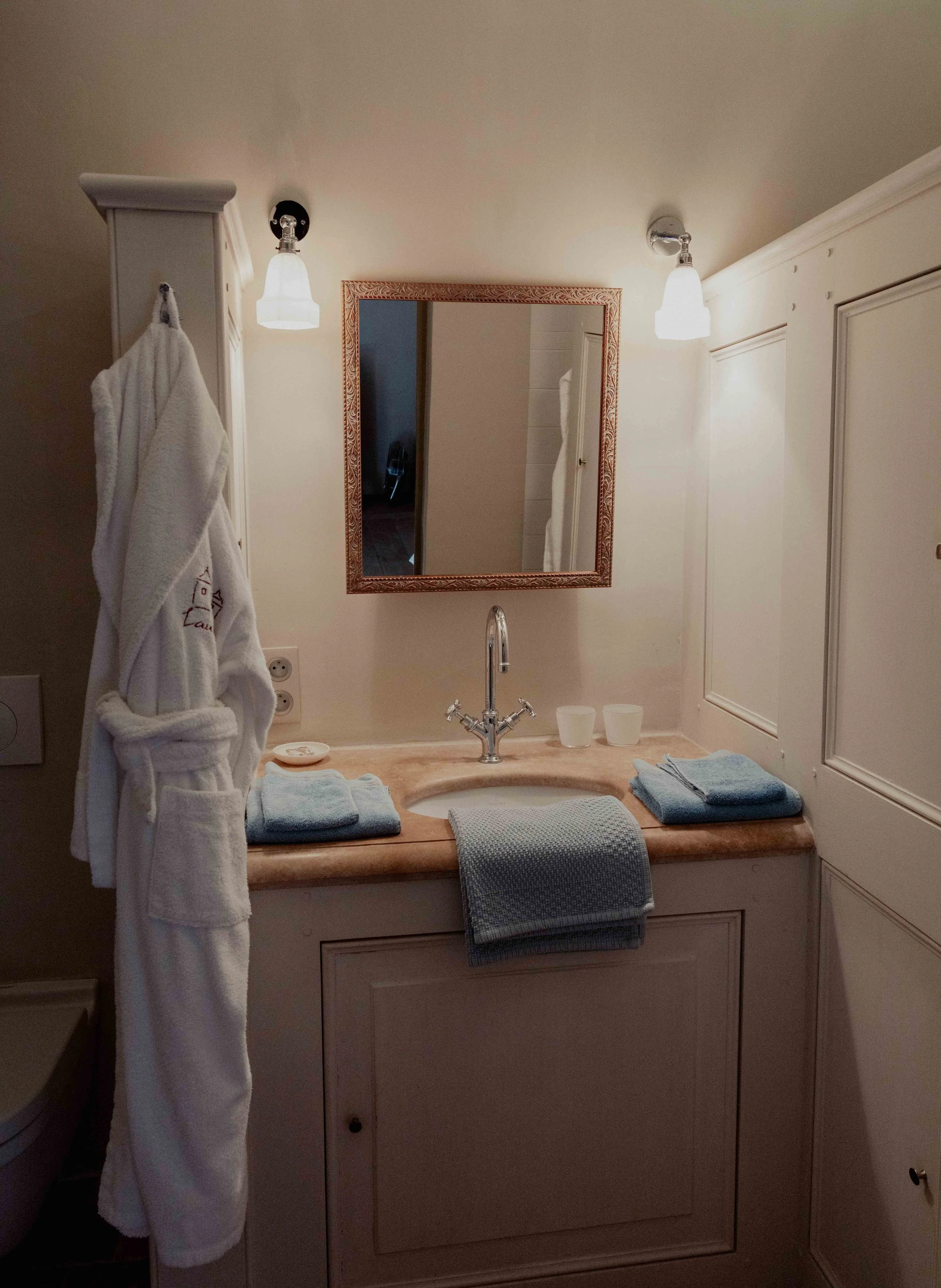 Een badkamer met een spiegel, wastafel, handdoeken en een badjas die aan een haak hangt.