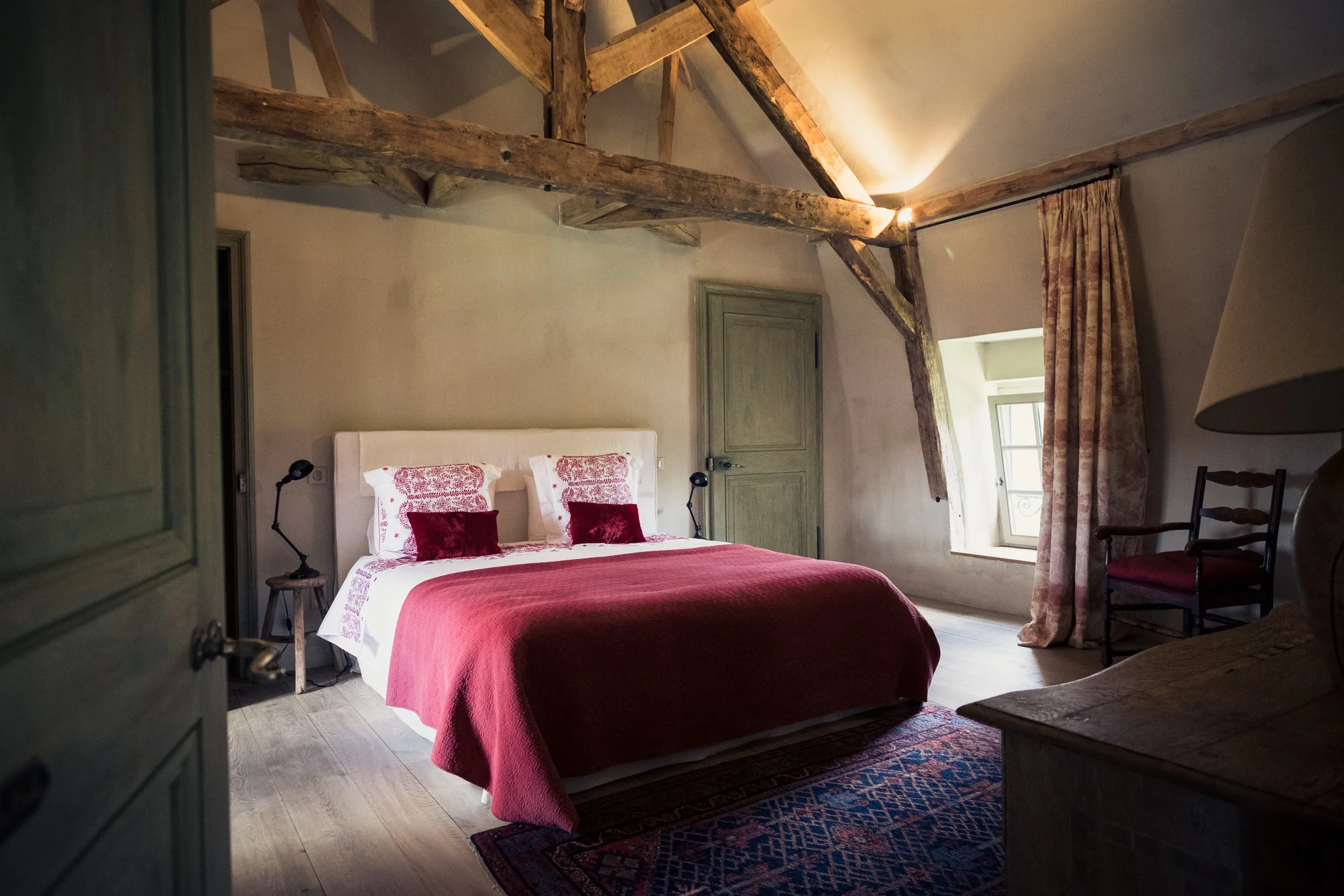A cozy rustic bedroom with a bed dressed in white and red bedding, rustic wooden beams on the ceiling, a window with floral curtains, and vintage furniture.